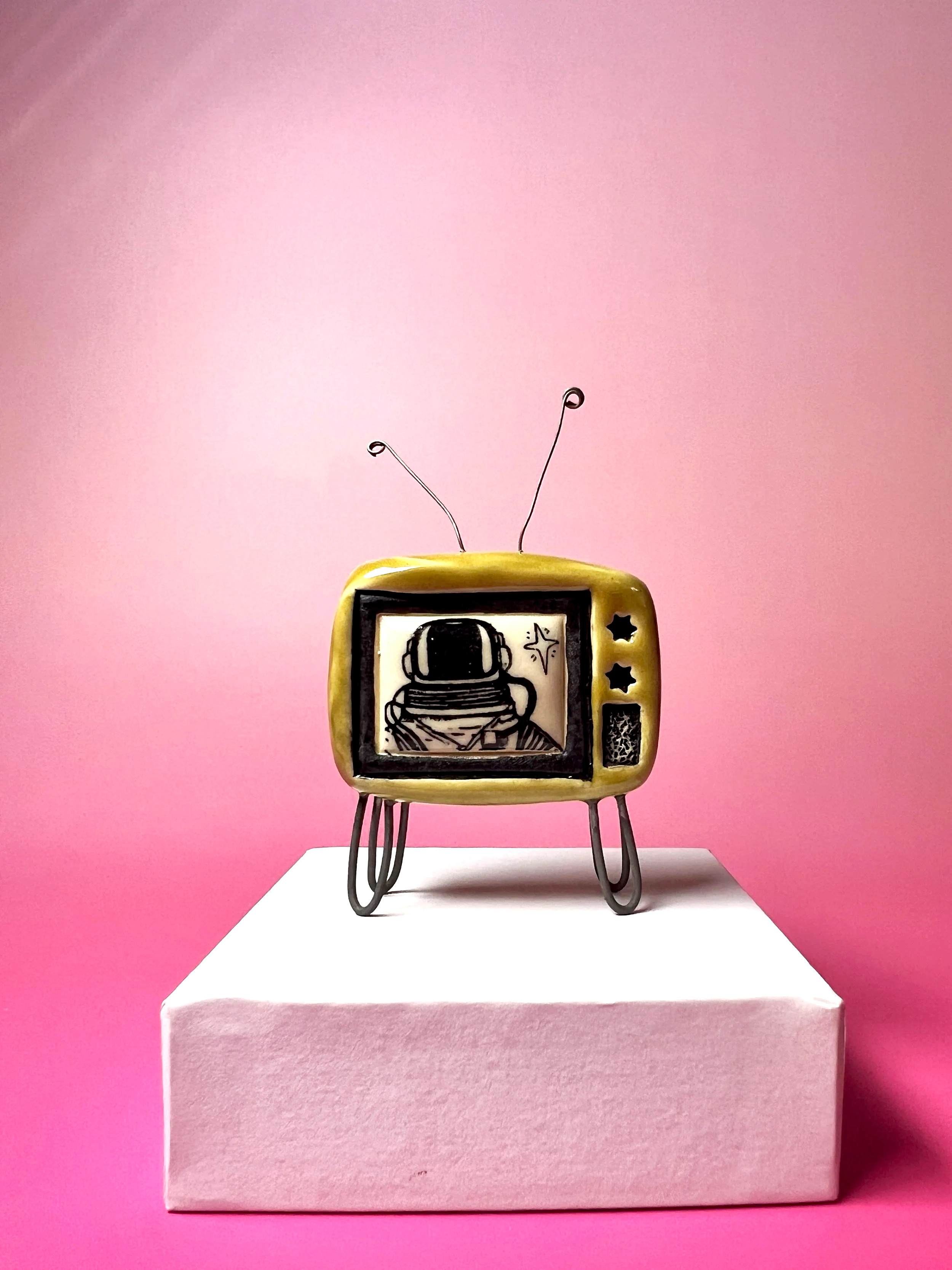 ceramic miniature TV with antenna and a scene of an astronaut on the screen on a white platform on a pink background