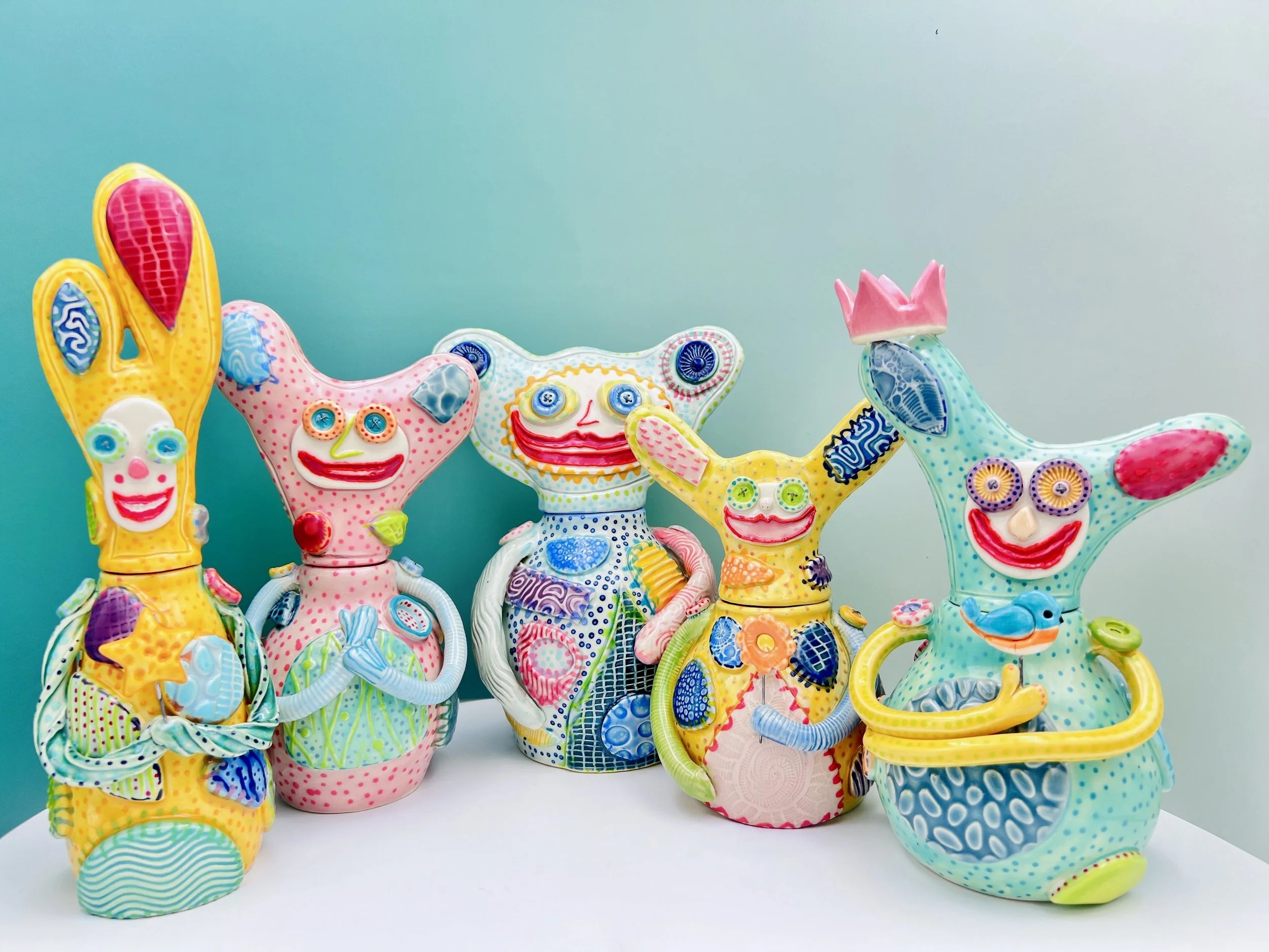 Colorful ceramic figurines with whimsical faces and detailing, arranged on a white surface against a blue gradient background.