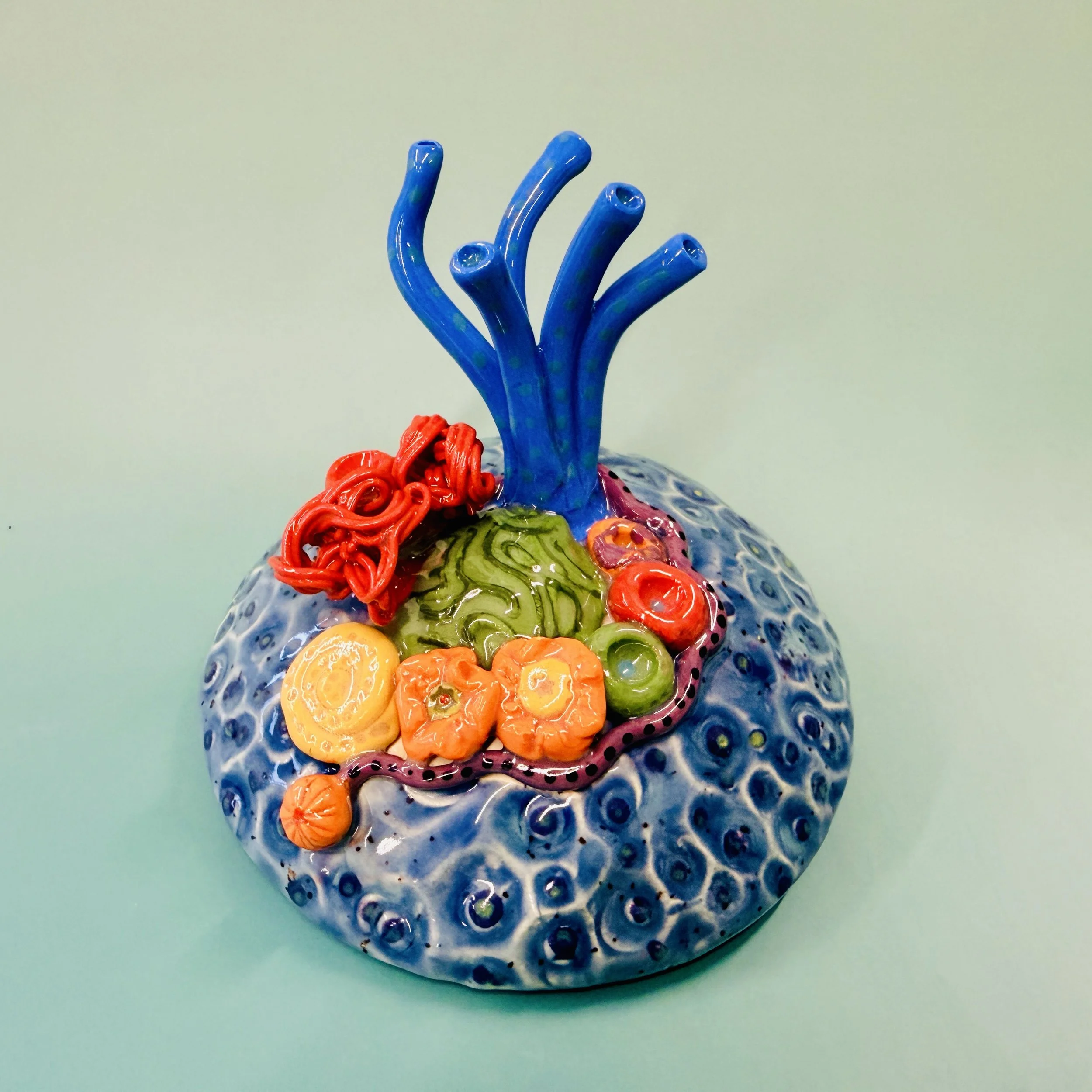 Colorful, textured clay sculptures resembling coral reefs arranged on a gradient background. Each piece has vibrant, intricate details, evoking a sense of ocean life.