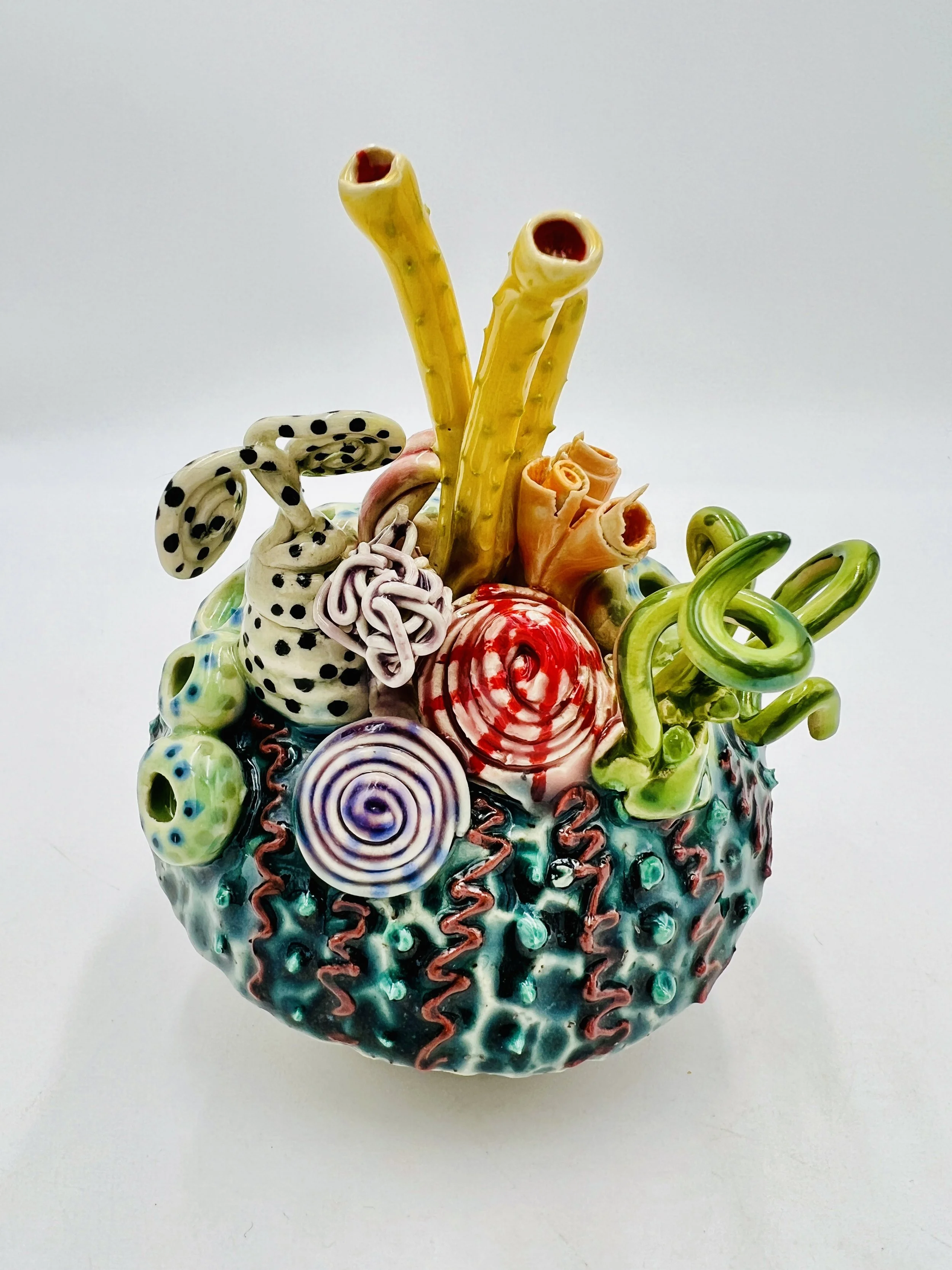 Colorful, textured clay sculptures resembling coral reefs arranged on a gradient background. Each piece has vibrant, intricate details, evoking a sense of ocean life.