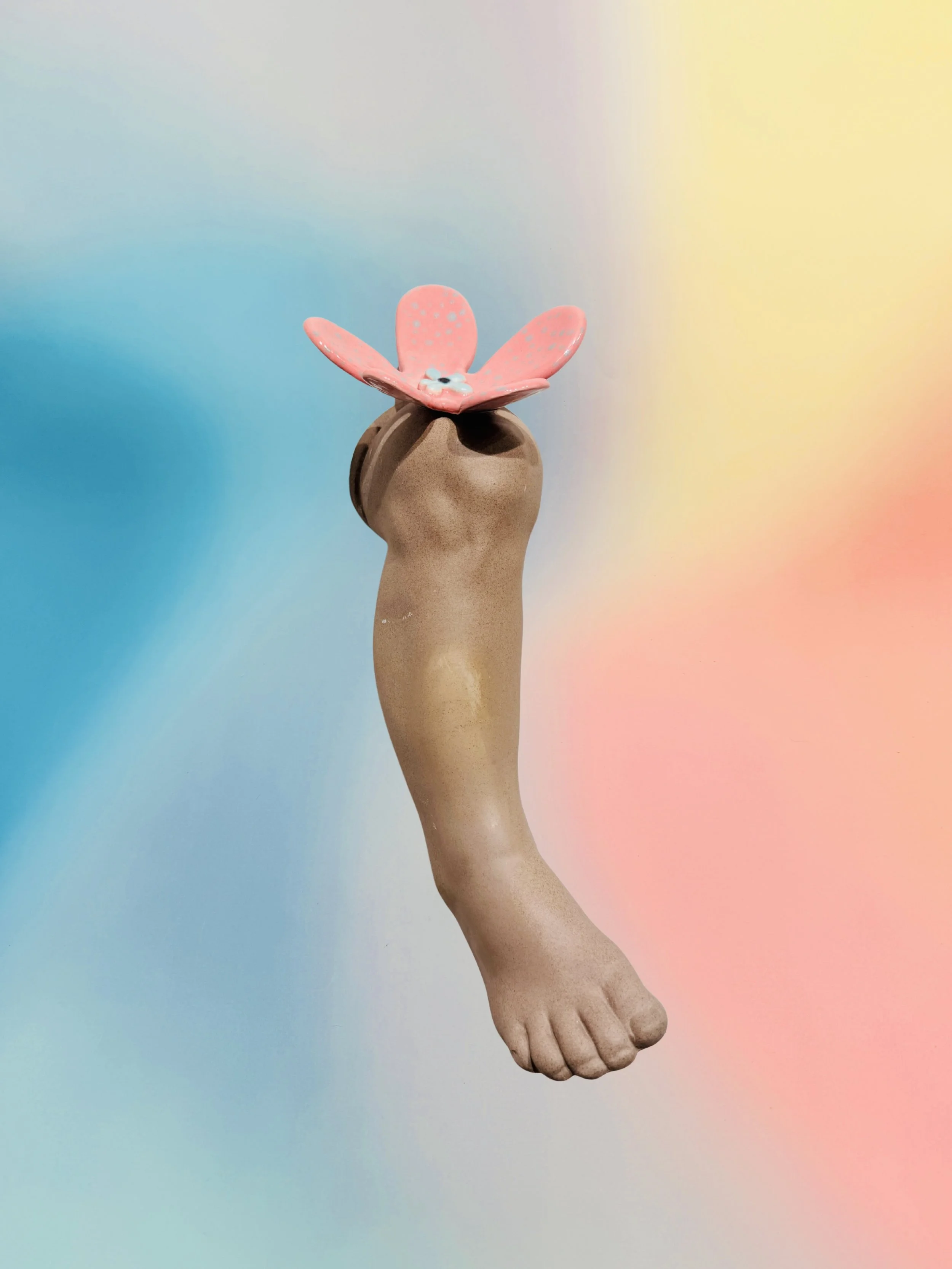 A tan sculptural leg hangs toes-down beneath a pink butterfly detail against a pastel blue, yellow, and coral backdrop, creating a surreal mood.