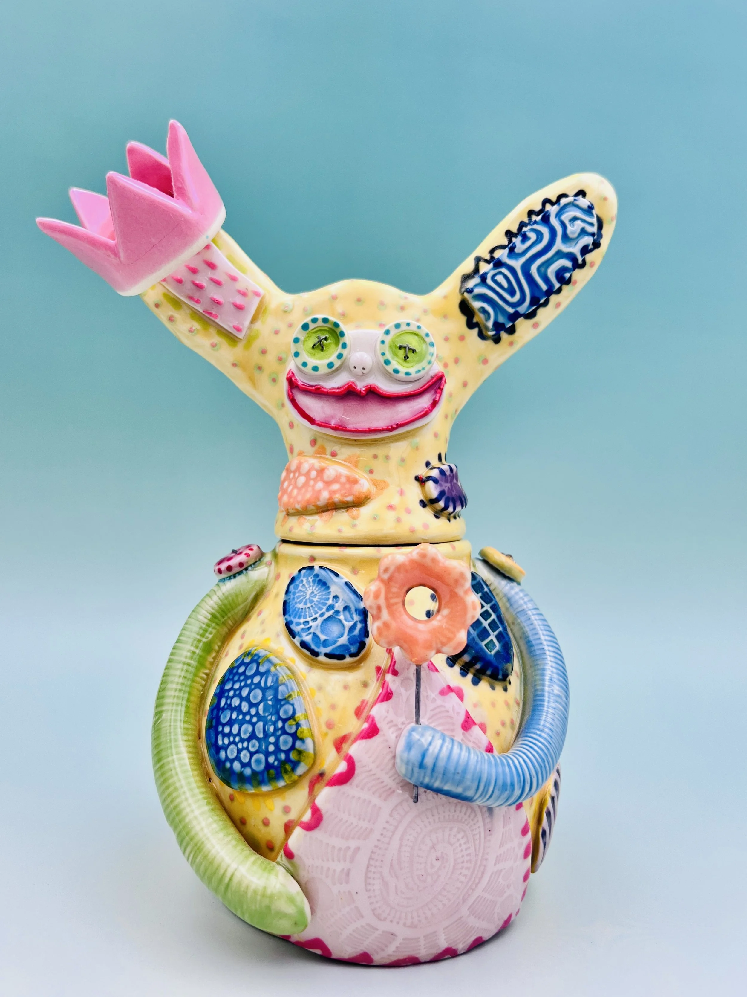 Colorful, whimsical ceramic figures called Gimmies with exaggerated facial features and various patterns, set against a gradient pastel background.