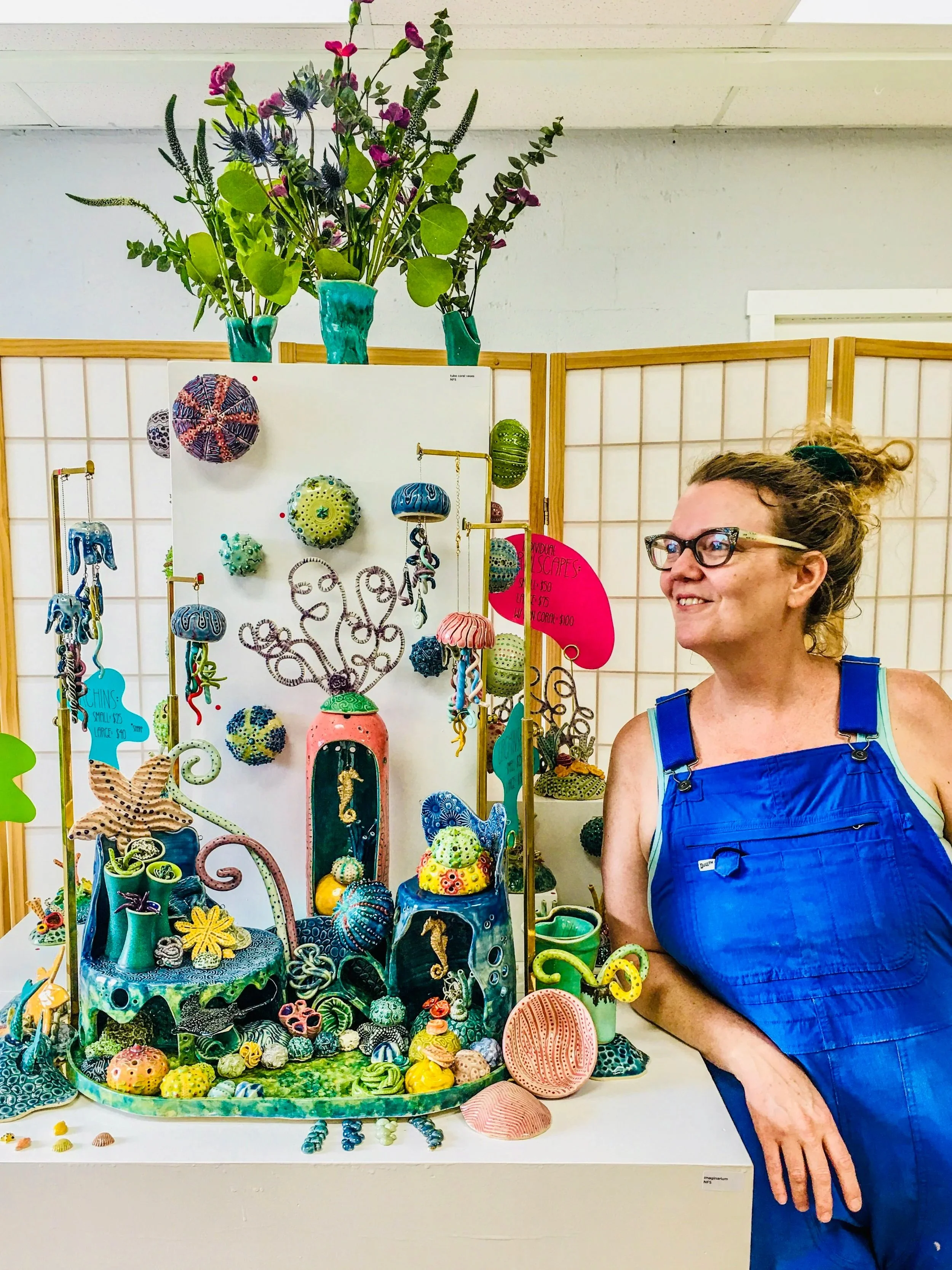 A woman with glasses and curly hair tied back, wearing a blue apron, standing next to a display of colorful, artistic sea life-themed sculptures and ornaments, including shells, sea creatures, and abstract designs, at an art or craft show.