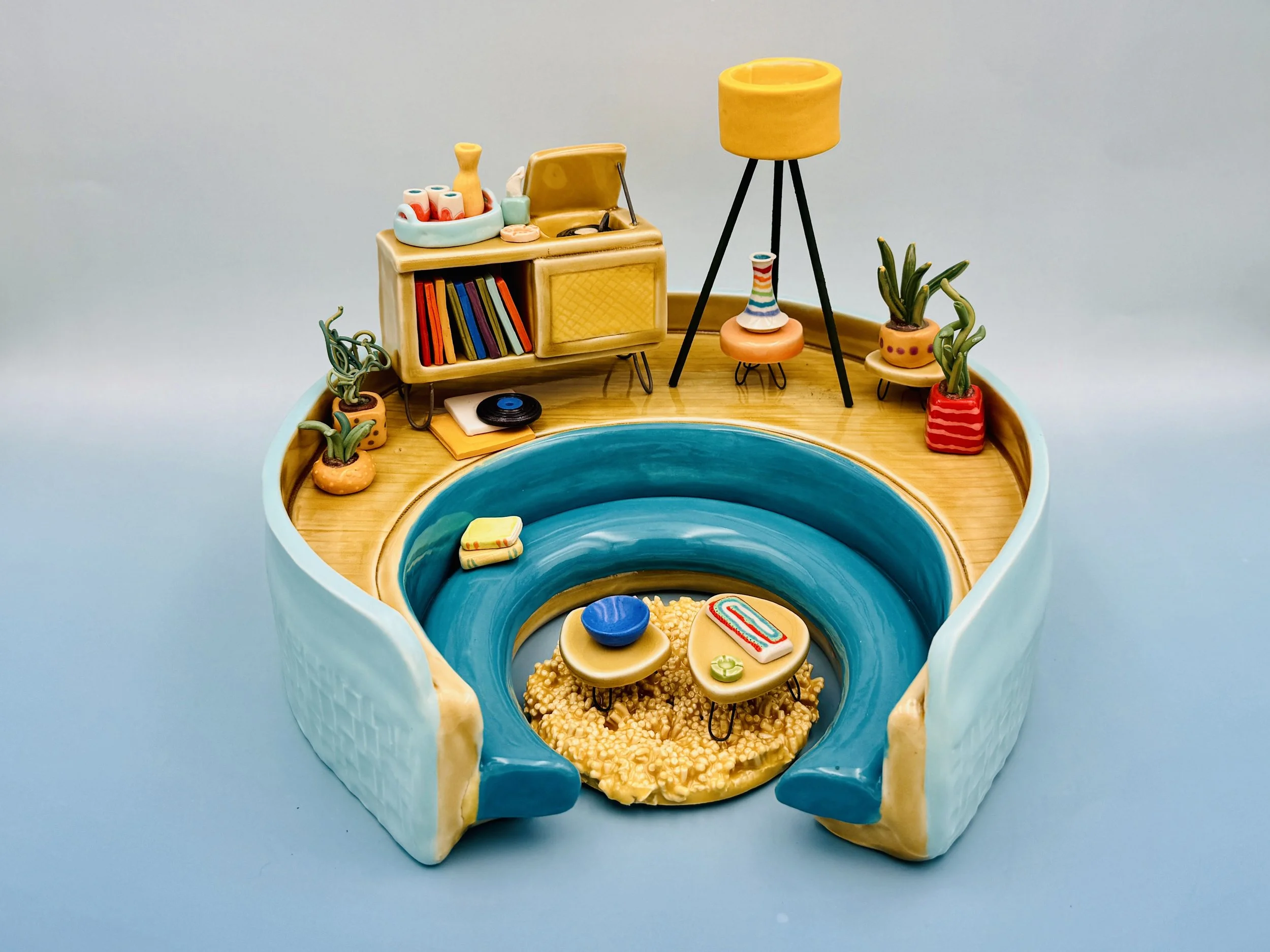 A colorful miniature room scene featuring blue curved benches, small tables with decorative items, a wooden cabinet with records, and various potted plants. A yellow floor lamp adds vibrant color.