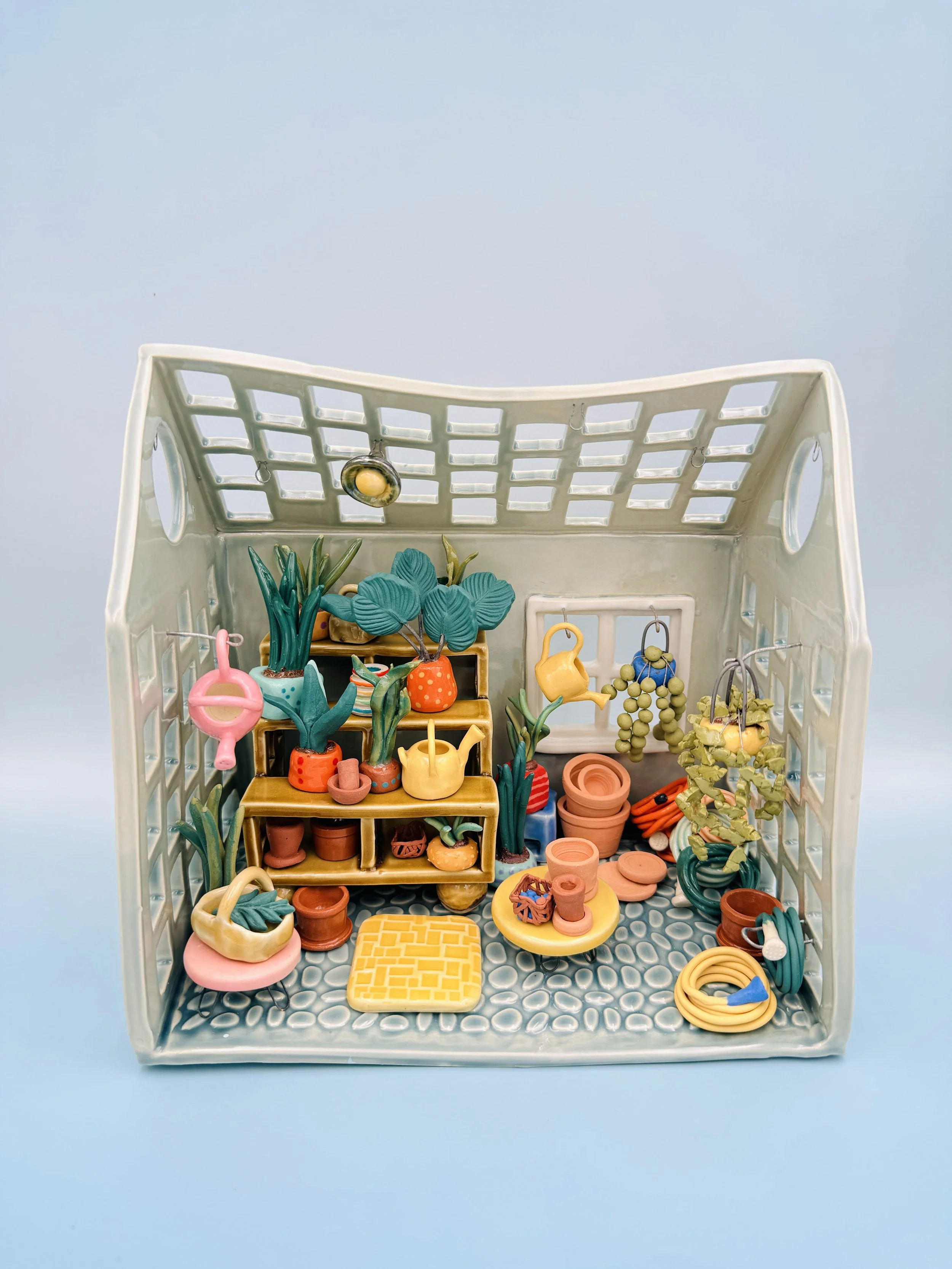The image showcases a ceramic whimsical indoor scene of a miniature garden space, presented in a structured container resembling a small house with a perforated roof and sides. The backdrop is a light blue, which contrasts with the vibrant colors of 