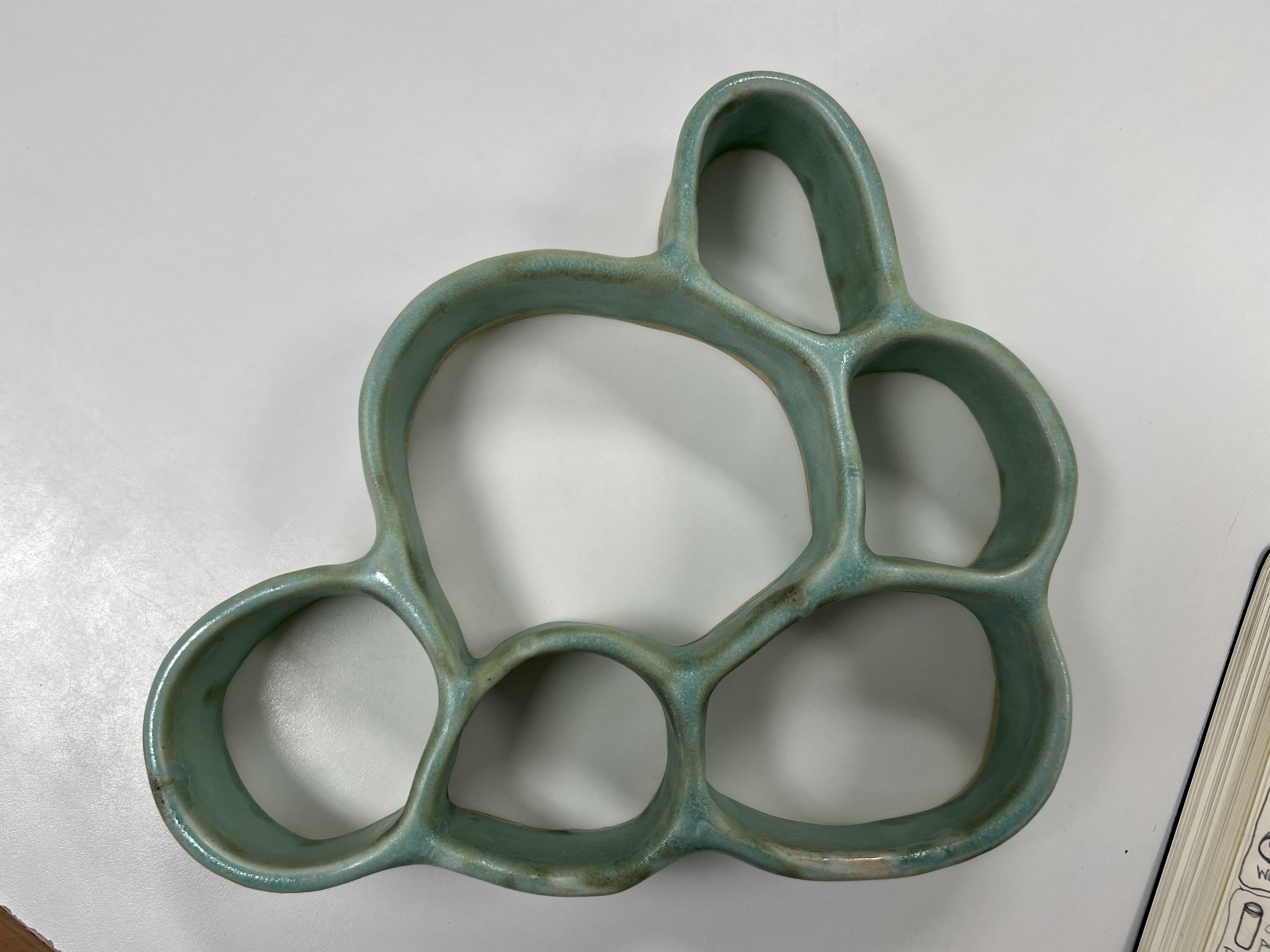 Green ceramic object with interconnected rounded loops, placed on a white surface with part of a notepad and a sheet of paper visible nearby.