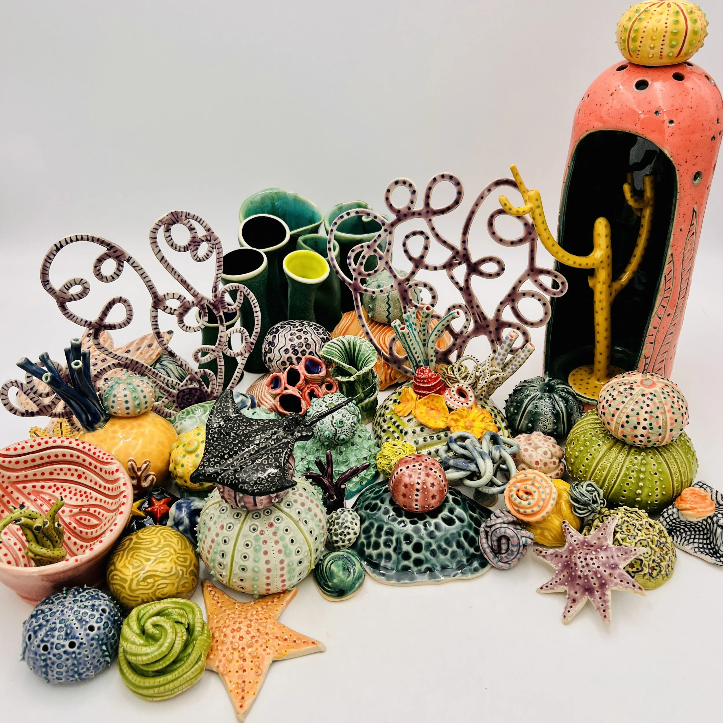 Colorful, textured clay sculptures resembling coral reefs arranged on a gradient background. Each piece has vibrant, intricate details, evoking a sense of ocean life.