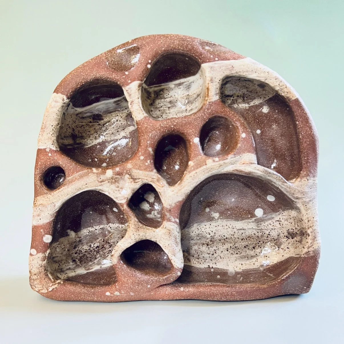 Abstract ceramic sculpture with smooth, rounded cavities. The earthy tones and irregular form evoke a natural, organic feel against a soft green background.