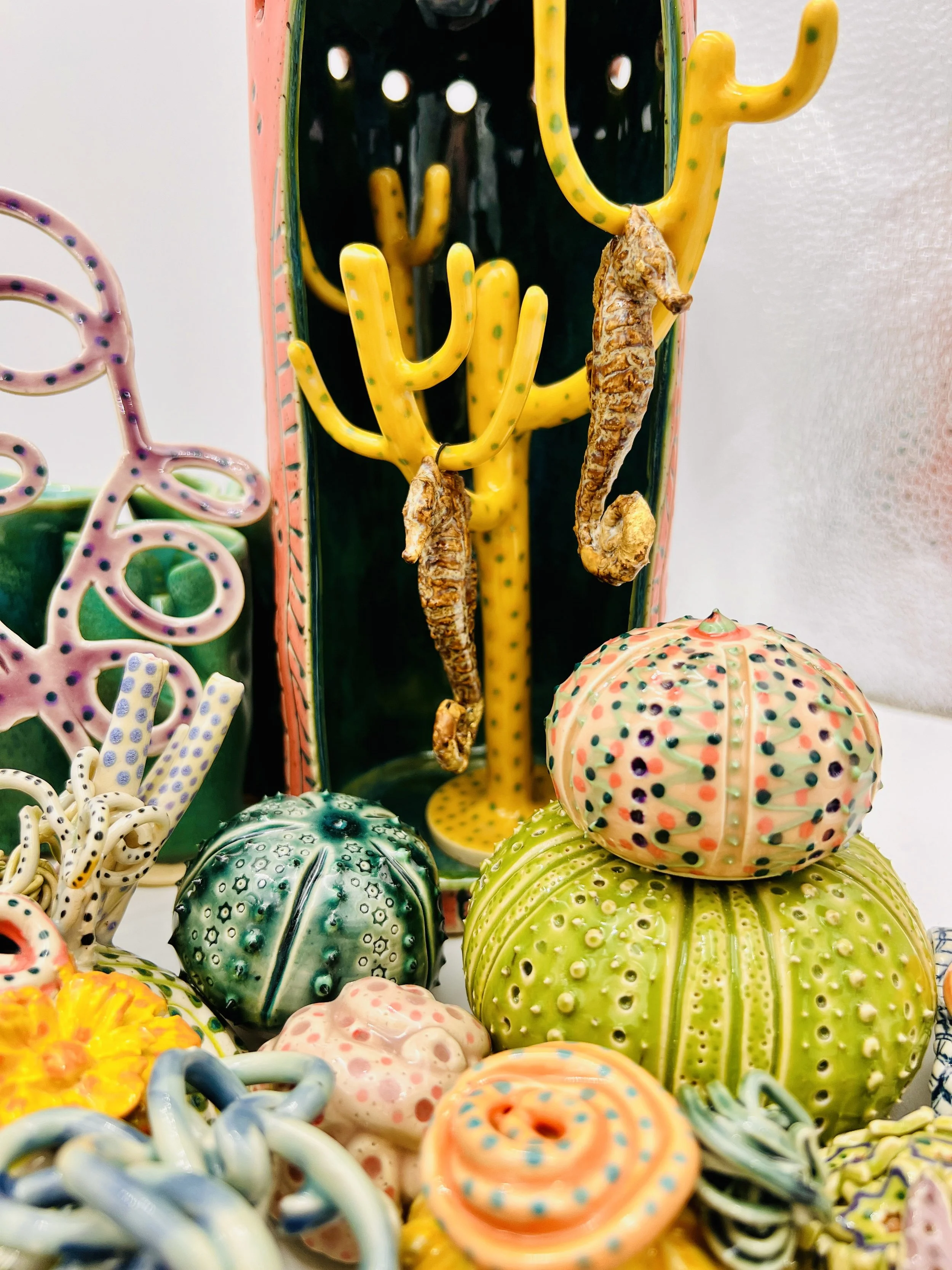 Colorful, textured clay sculptures resembling coral reefs arranged on a gradient background. Each piece has vibrant, intricate details, evoking a sense of ocean life.