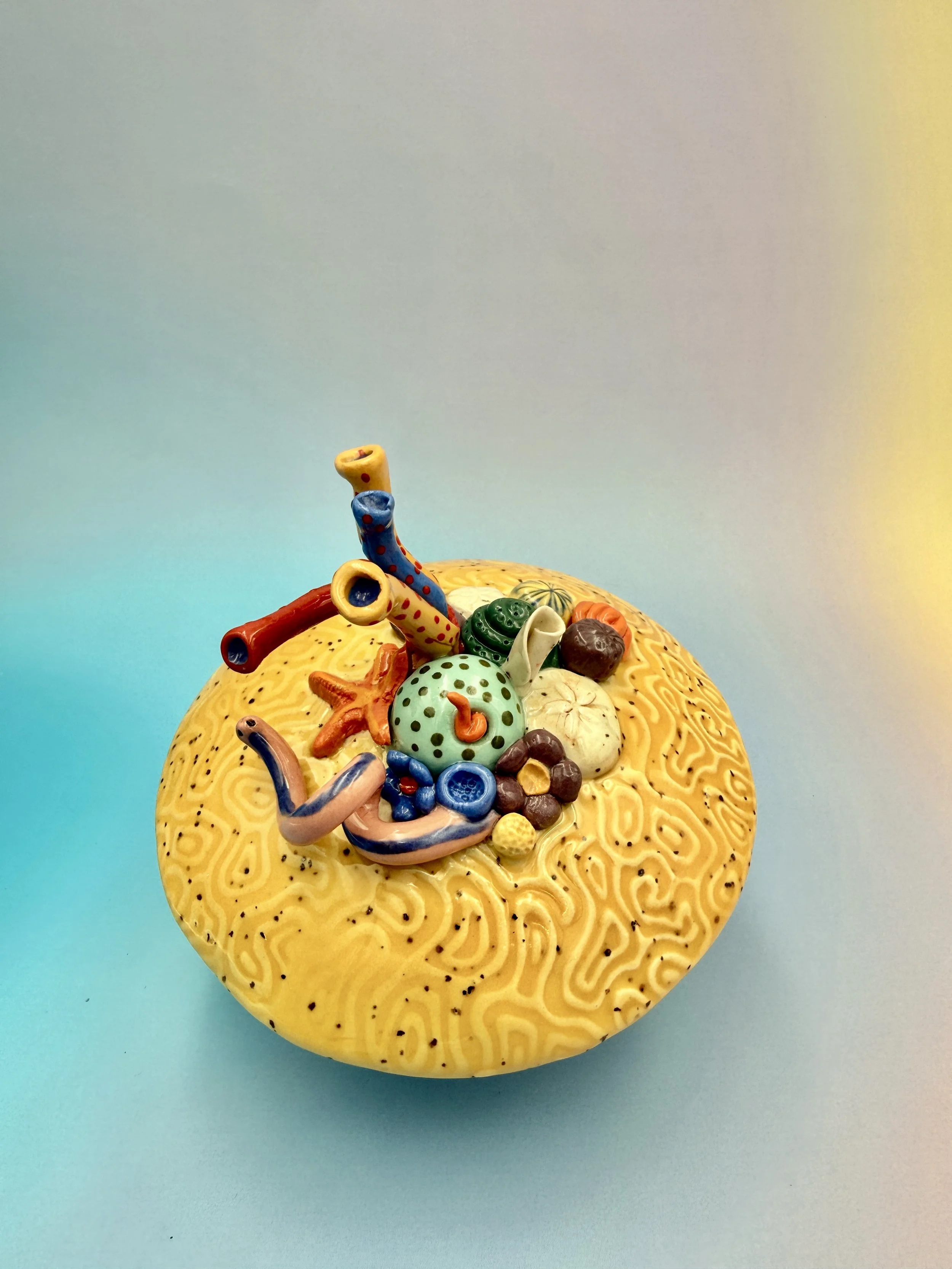 Colorful, textured clay sculptures resembling coral reefs arranged on a gradient background. Each piece has vibrant, intricate details, evoking a sense of ocean life.
