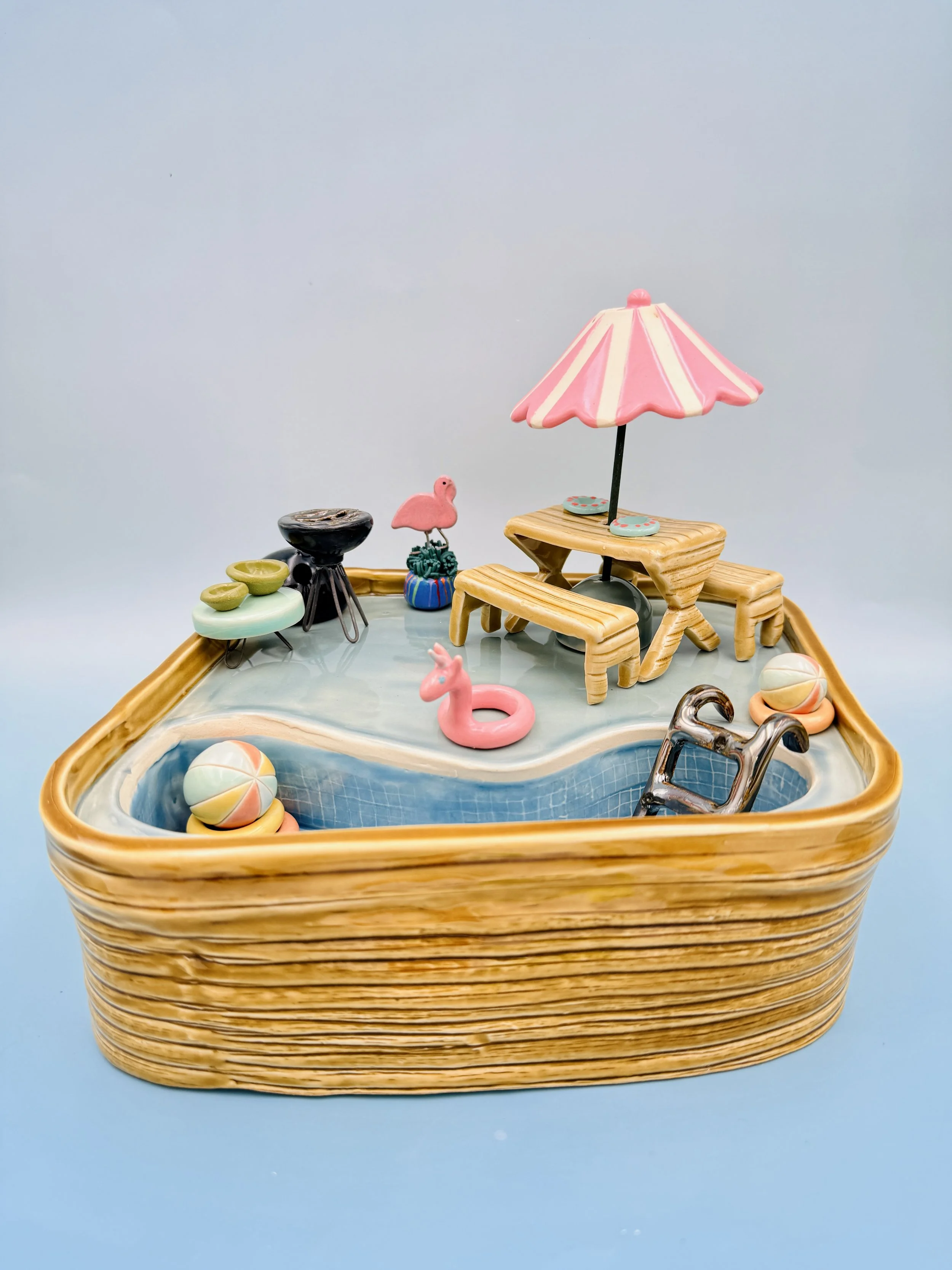 The image depicts a small, playful ceramic model of a backyard pool scene. The pool is shaped irregularly and features a light blue surface with a textured bottom that suggests water. Surrounding the pool is a tan-colored, textured rim resembling wic