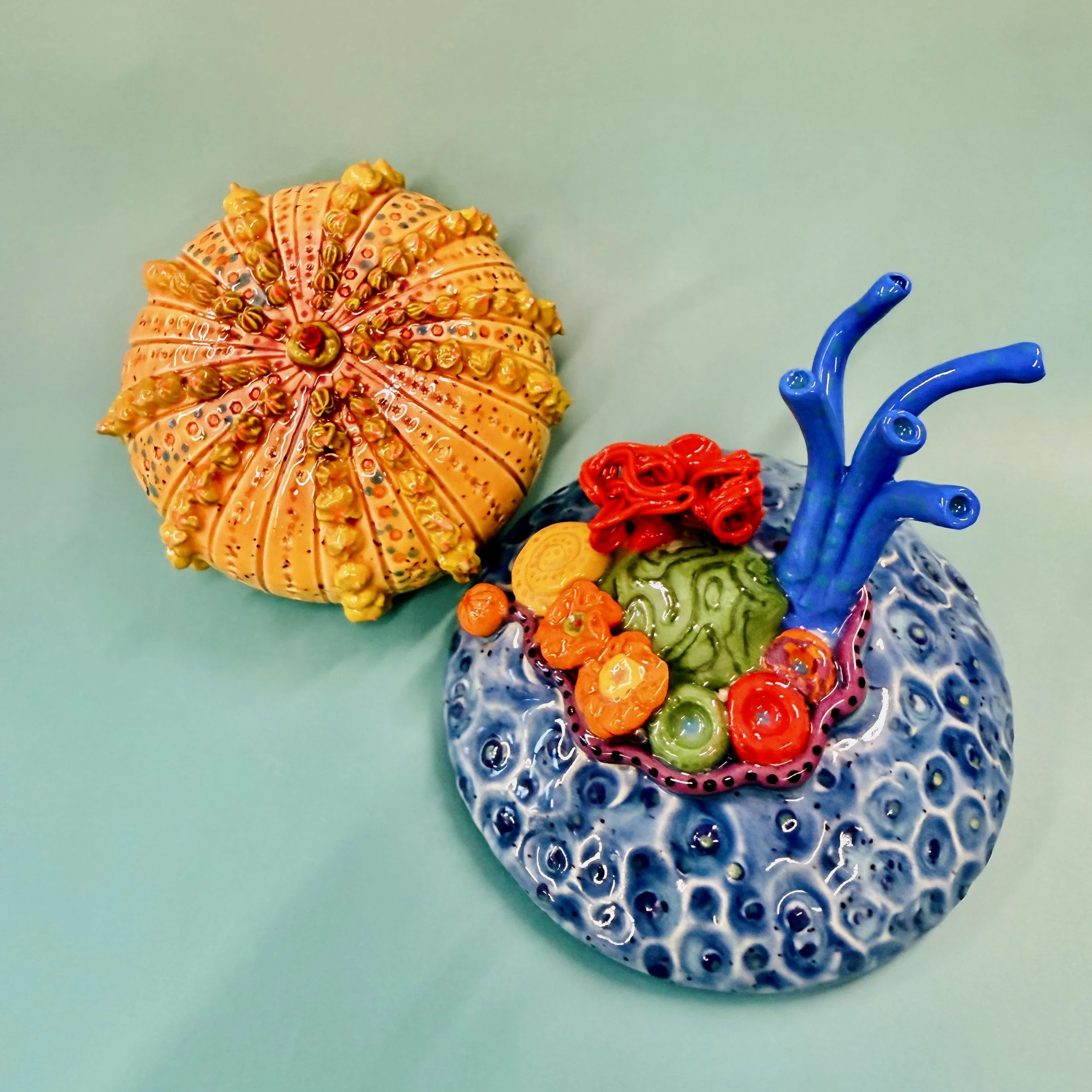 Colorful, textured clay sculptures resembling coral reefs arranged on a gradient background. Each piece has vibrant, intricate details, evoking a sense of ocean life.