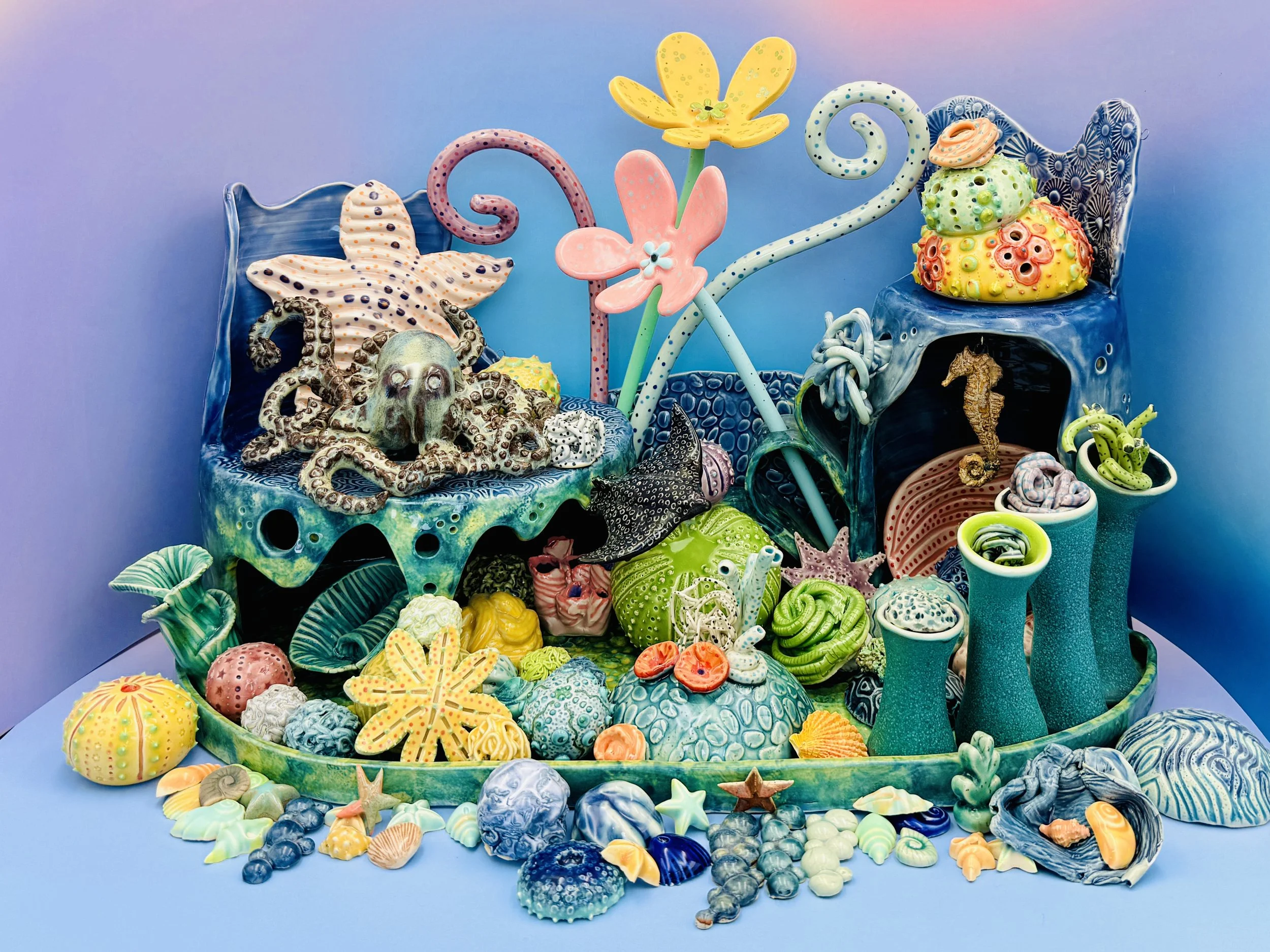 The image portrays an intricate underwater scene crafted from vibrant pottery, resembling a colorful coral reef. In the center, a detailed model of an octopus is nestled among green and blue corals, showcasing its textured body and large eyes. Surrou