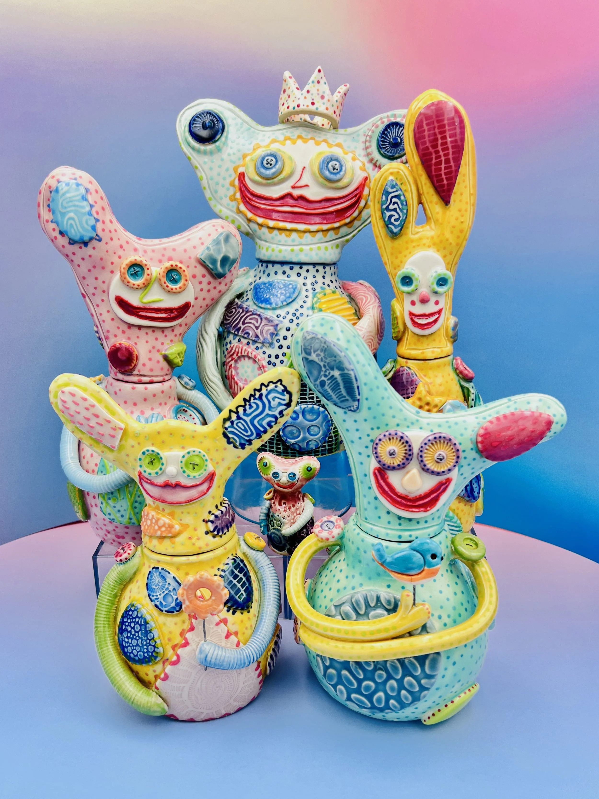 Colorful, whimsical ceramic figures called Gimmies with exaggerated facial features and various patterns, set against a gradient pastel background.