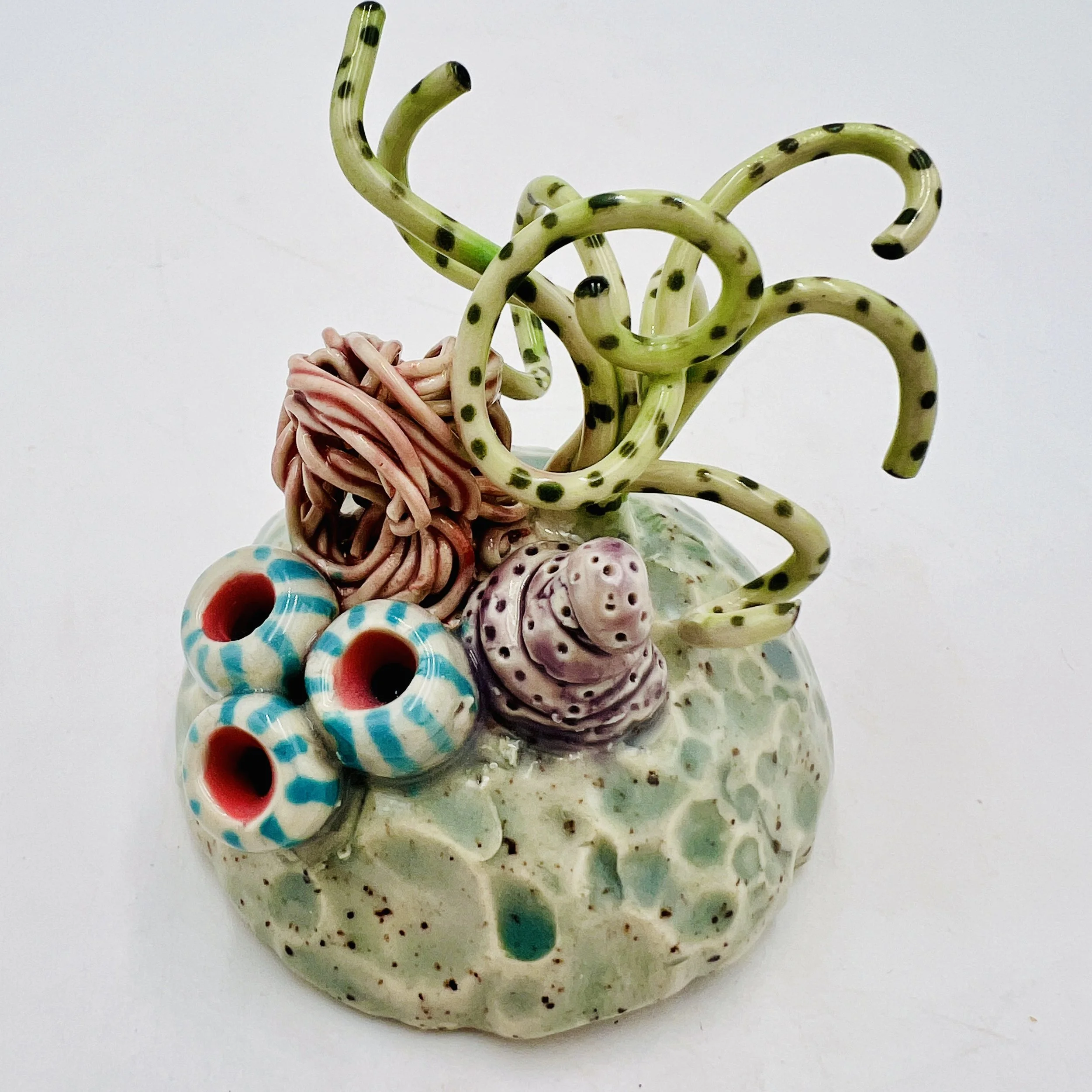 Colorful, textured clay sculptures resembling coral reefs arranged on a gradient background. Each piece has vibrant, intricate details, evoking a sense of ocean life.