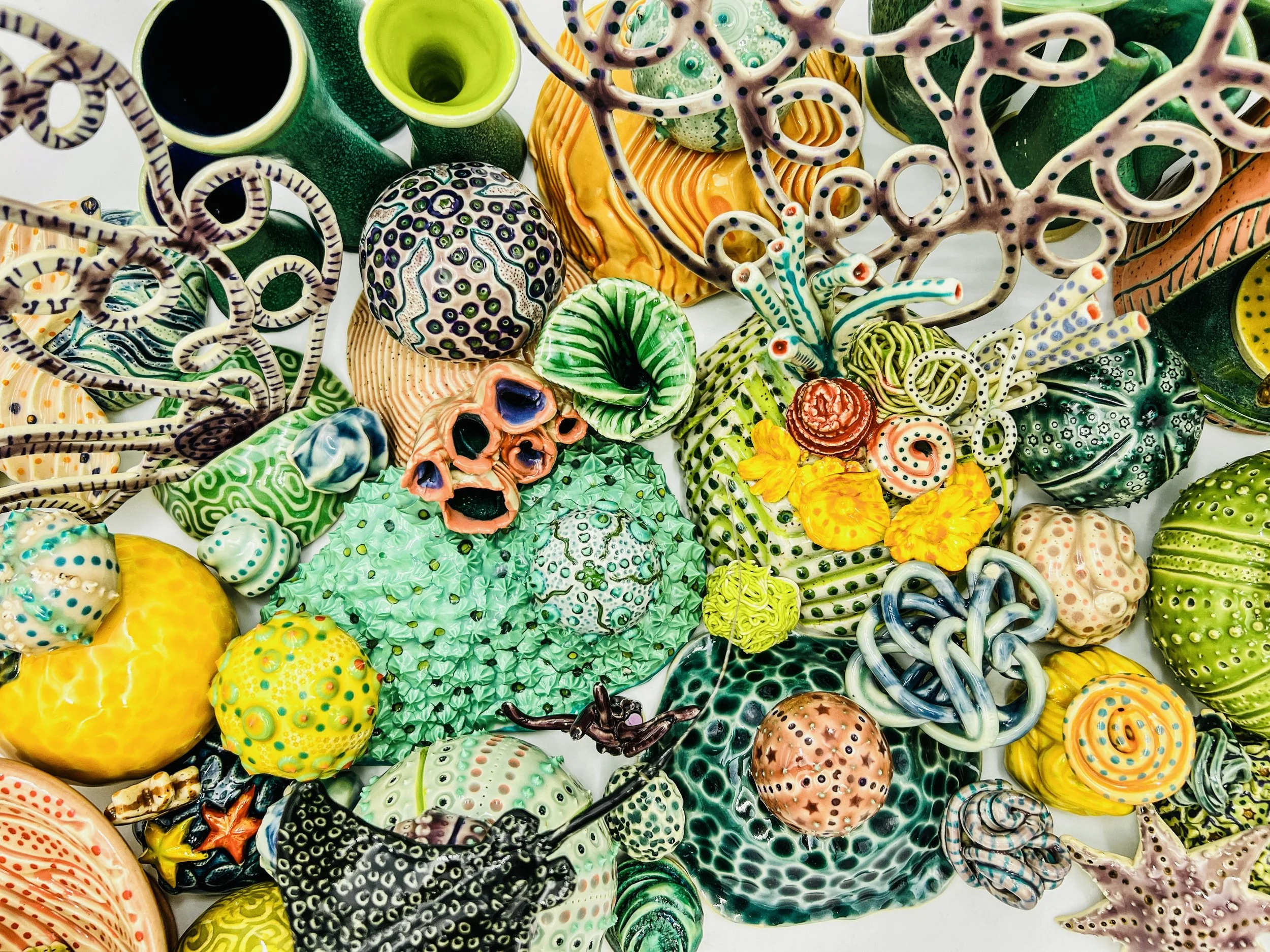 Colorful, textured clay sculptures resembling coral reefs arranged on a gradient background. Each piece has vibrant, intricate details, evoking a sense of ocean life.