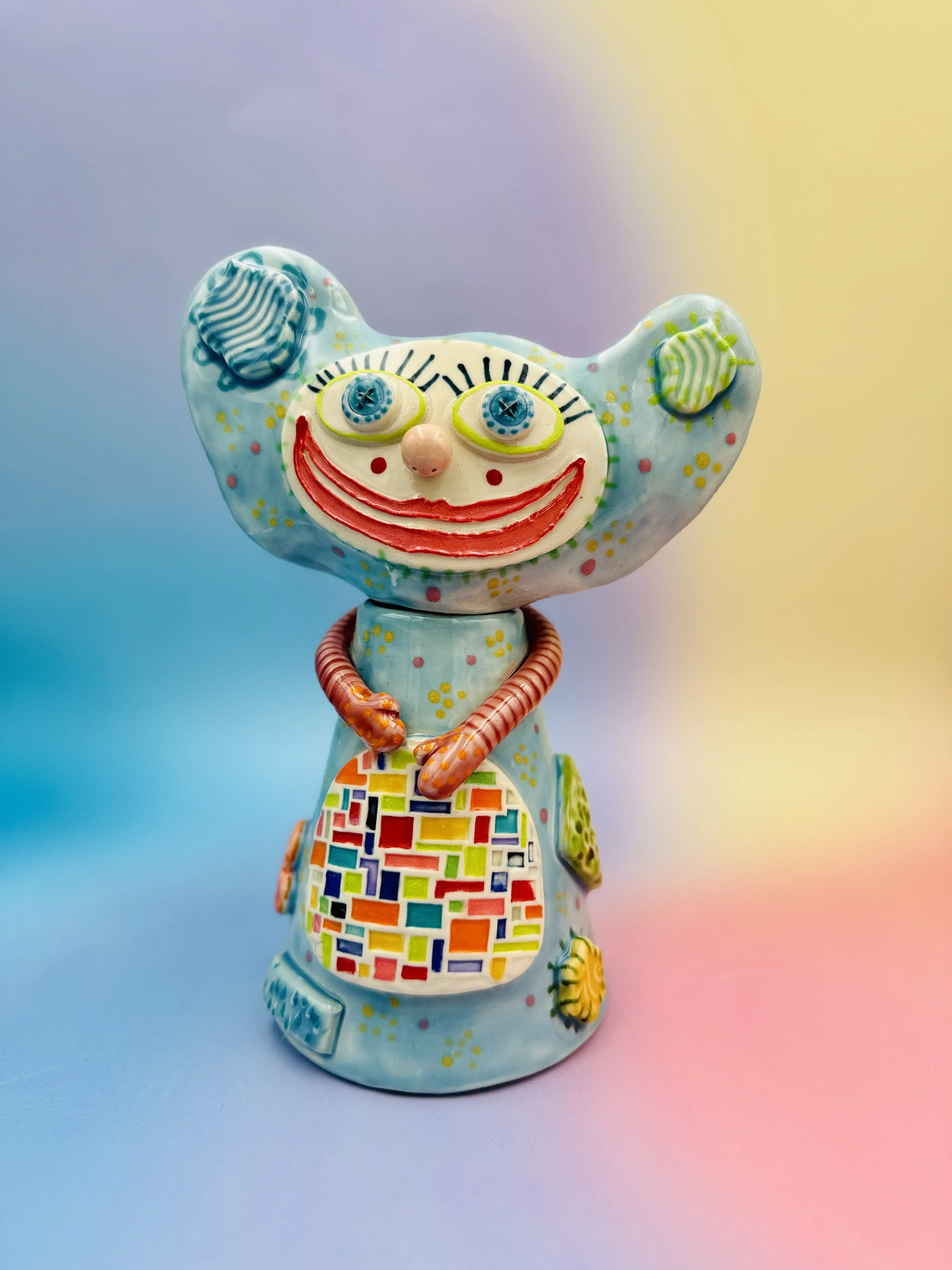 Colorful, whimsical ceramic figures called Gimmies with exaggerated facial features and various patterns, set against a gradient pastel background.