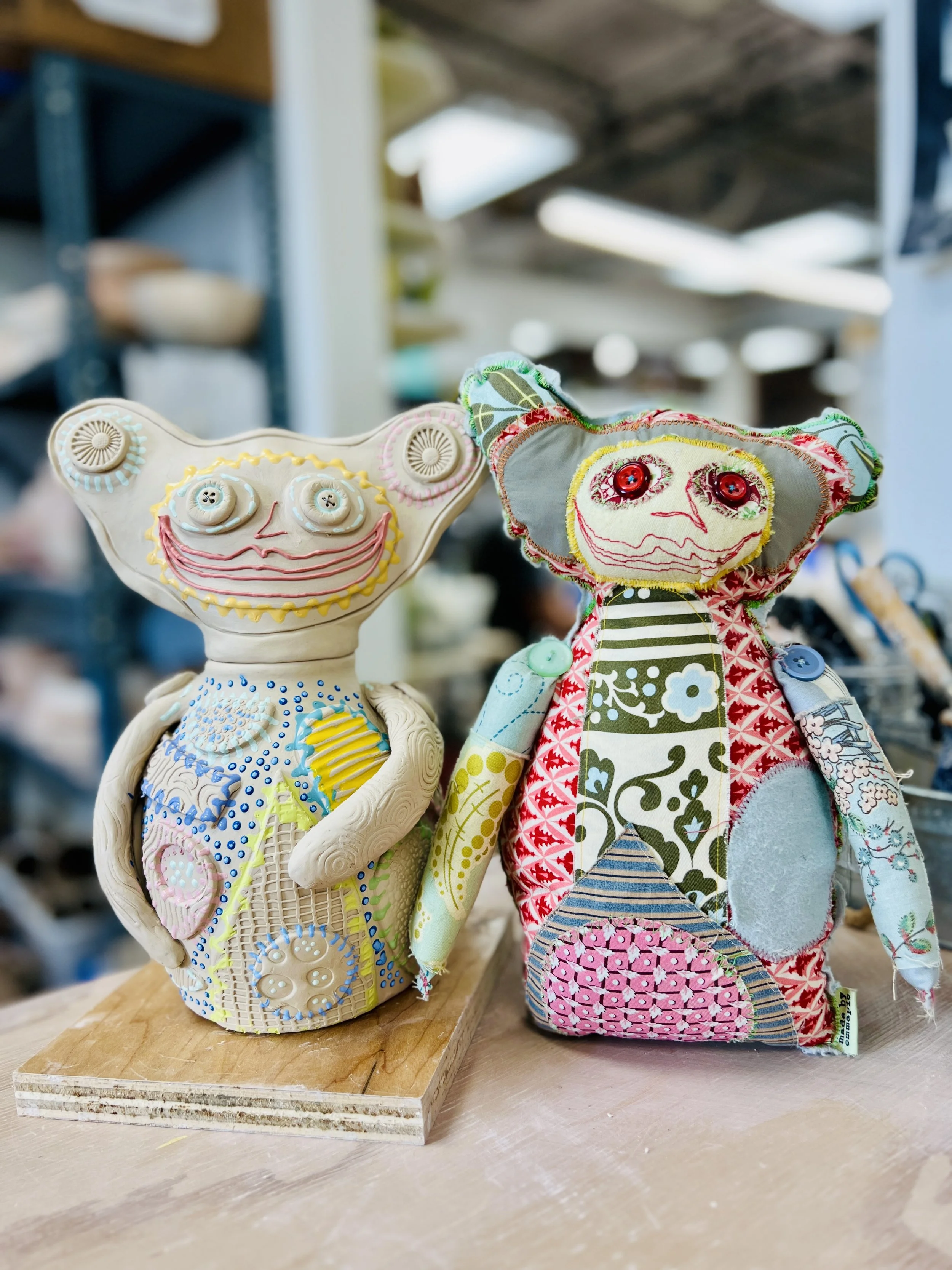 Colorful, whimsical ceramic figures called Gimmies with exaggerated facial features and various patterns next to a sewn version of one