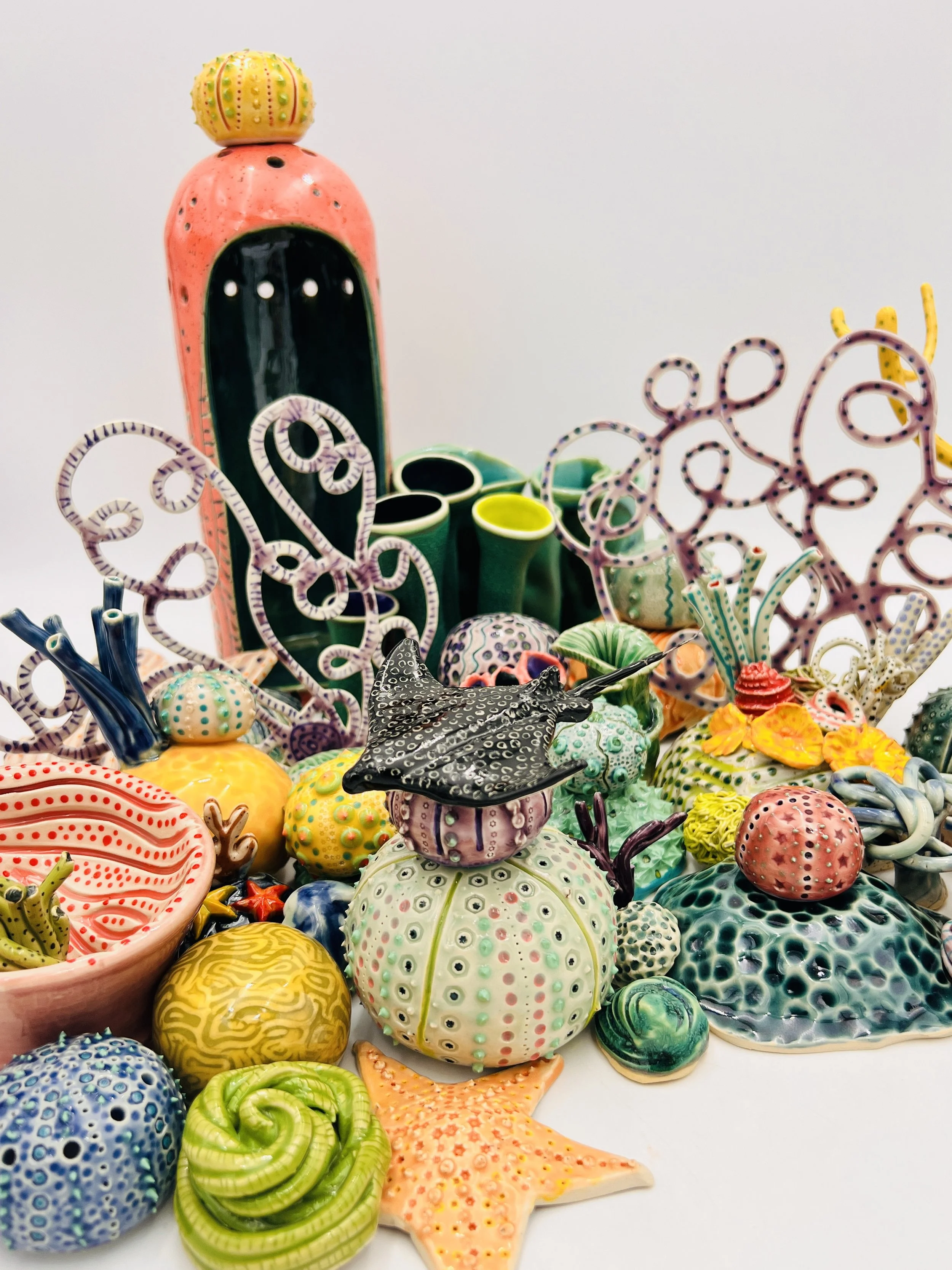 Colorful, textured clay sculptures resembling coral reefs arranged on a gradient background. Each piece has vibrant, intricate details, evoking a sense of ocean life.