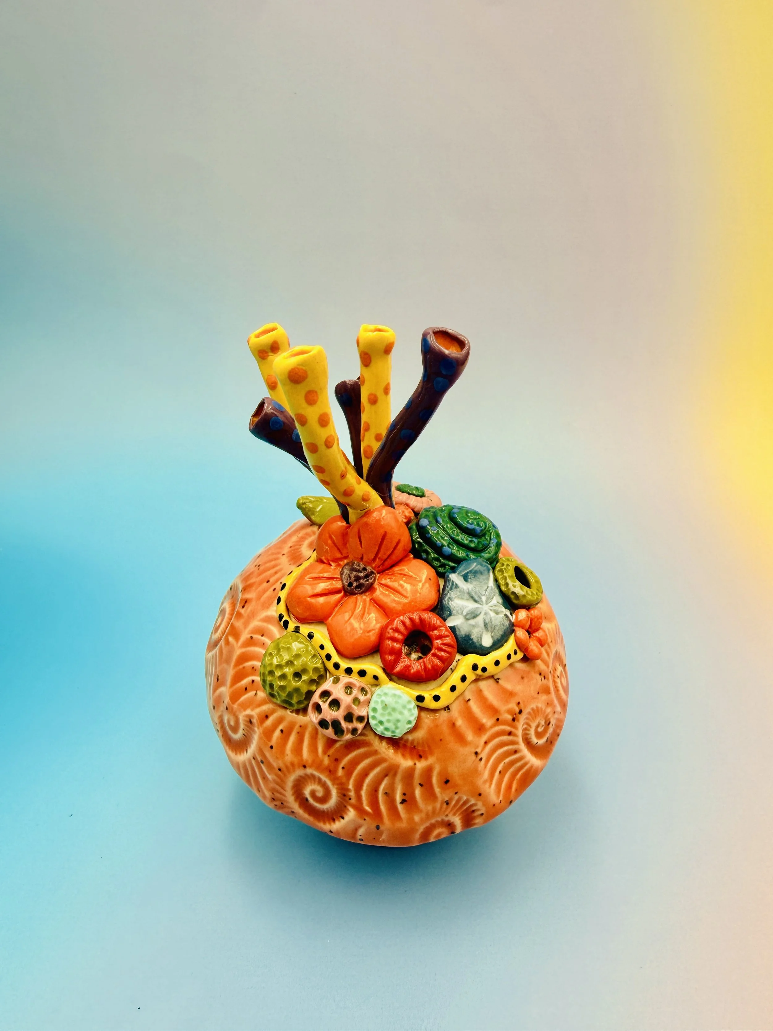 Colorful, textured clay sculptures resembling coral reefs arranged on a gradient background. Each piece has vibrant, intricate details, evoking a sense of ocean life.