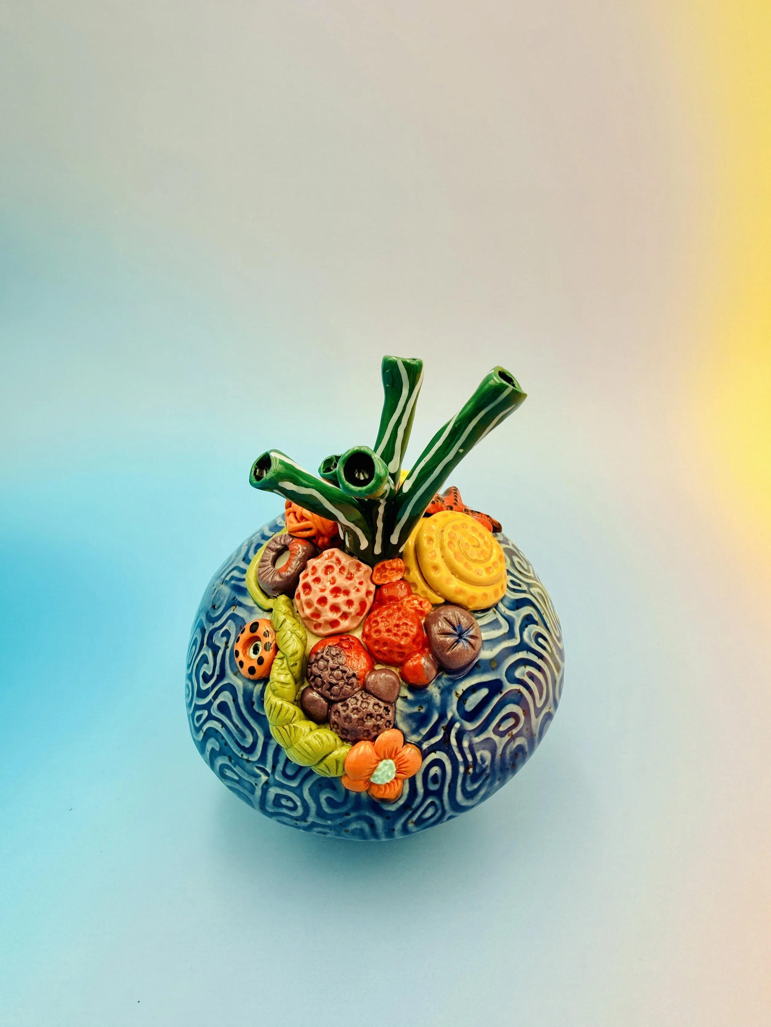 Colorful, textured clay sculptures resembling coral reefs arranged on a gradient background. Each piece has vibrant, intricate details, evoking a sense of ocean life.