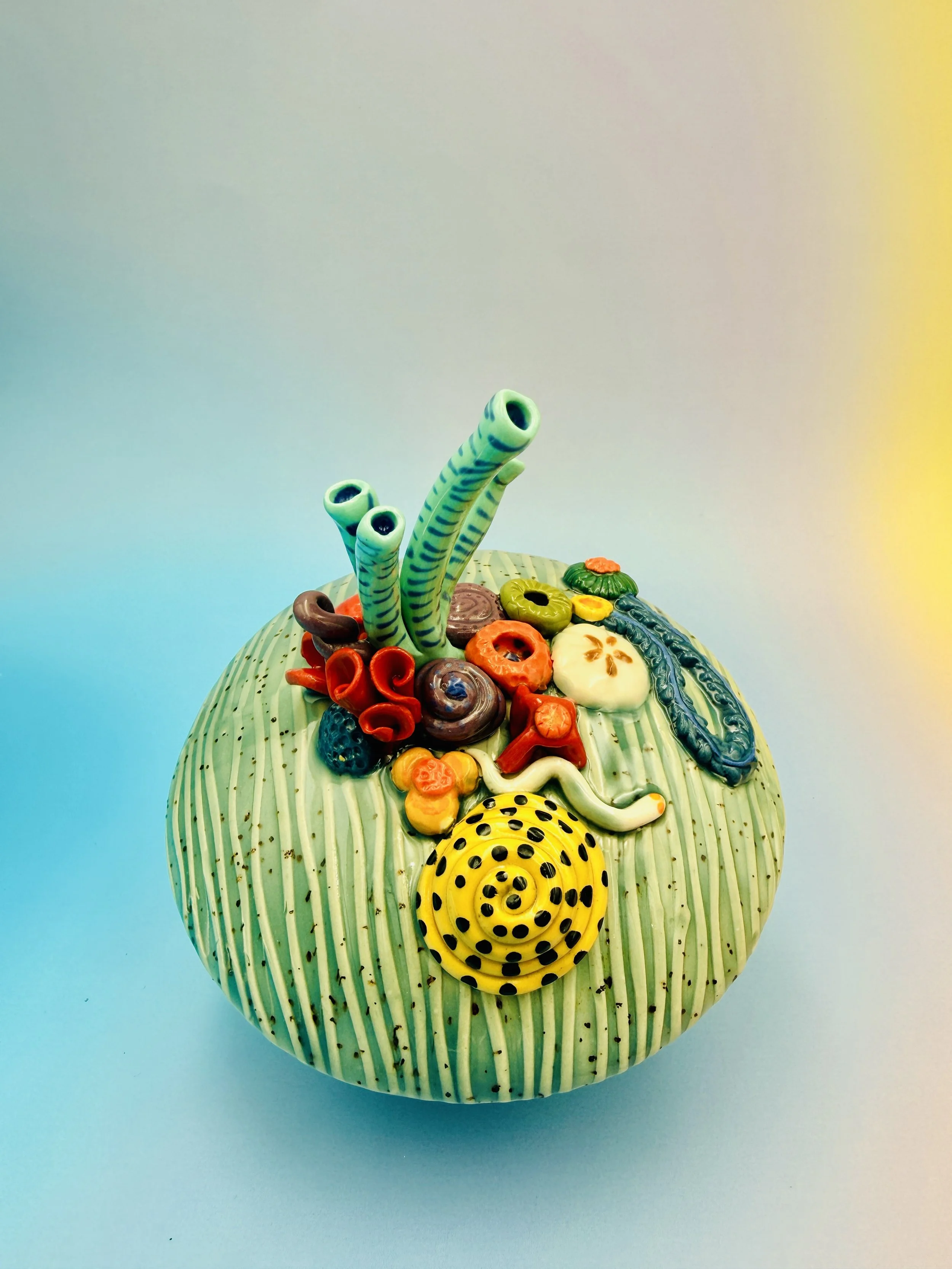 Colorful, textured clay sculptures resembling coral reefs arranged on a gradient background. Each piece has vibrant, intricate details, evoking a sense of ocean life.