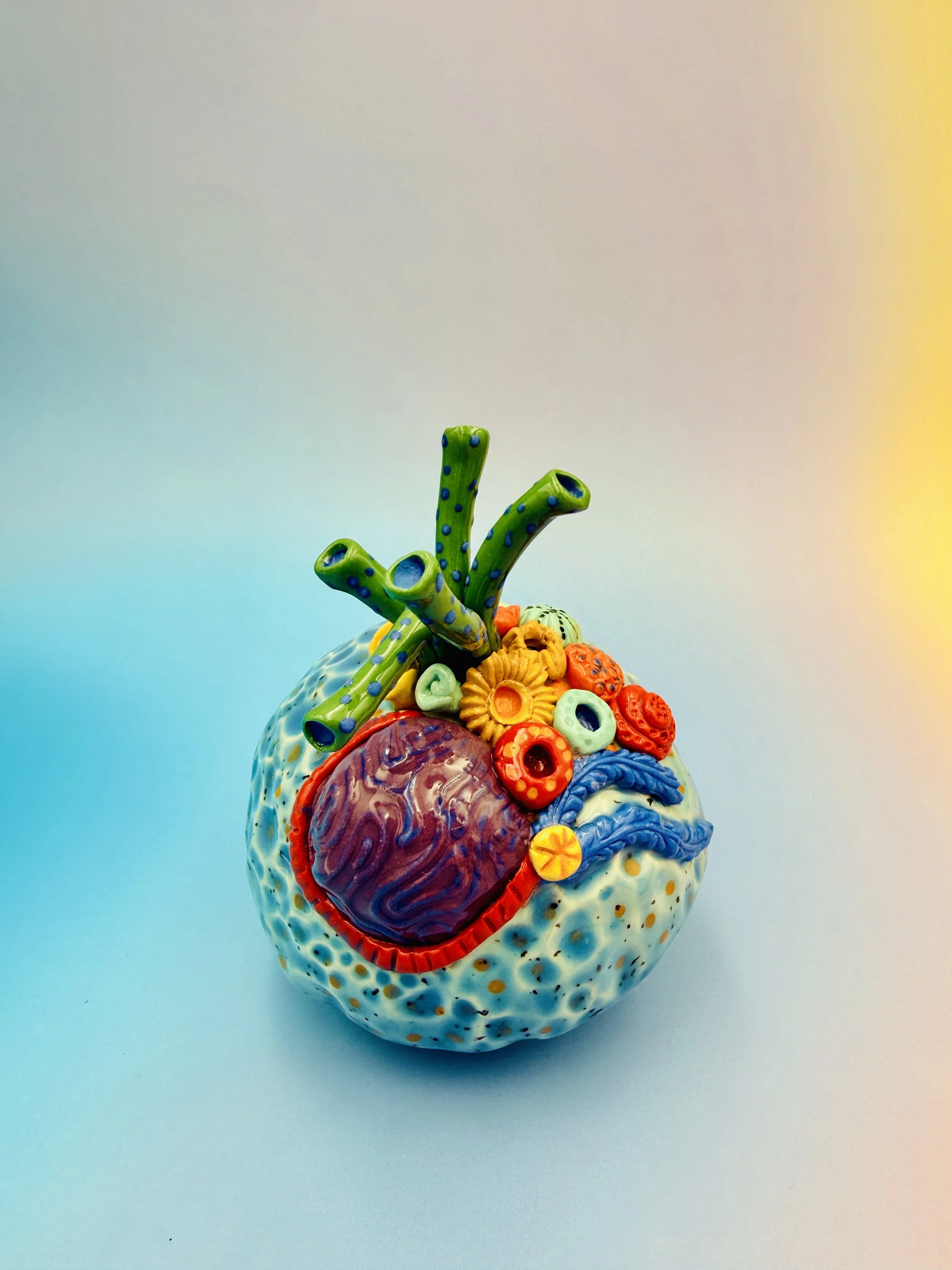 Colorful, textured clay sculptures resembling coral reefs arranged on a gradient background. Each piece has vibrant, intricate details, evoking a sense of ocean life.