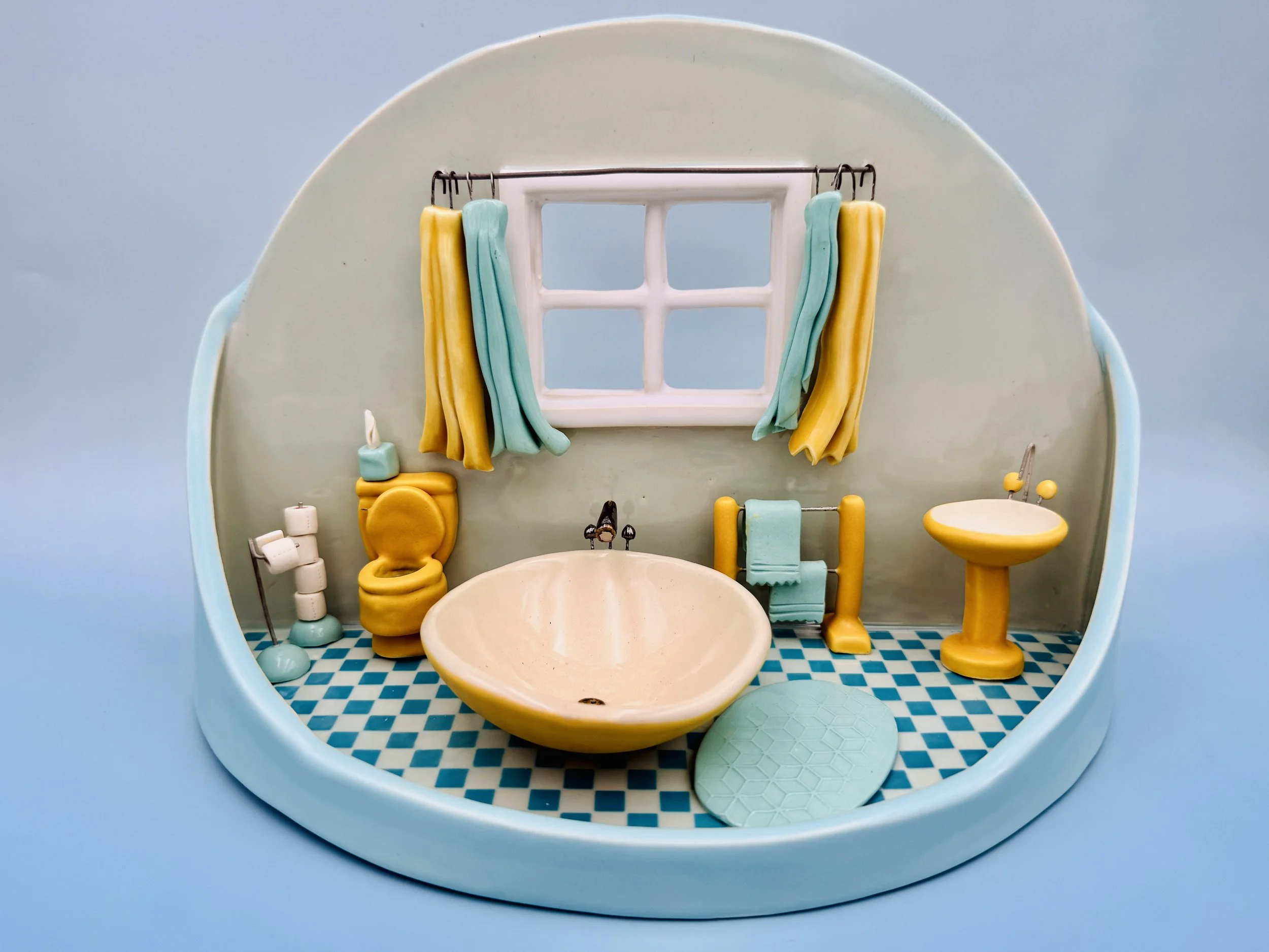 The image features a colorful ceramic miniature representation of a bathroom scene. The backdrop is a soft blue, enhancing the vibrant colors in the setup. On the left side, a yellow-and-aqua patterned shower curtain hangs beside a small window with 