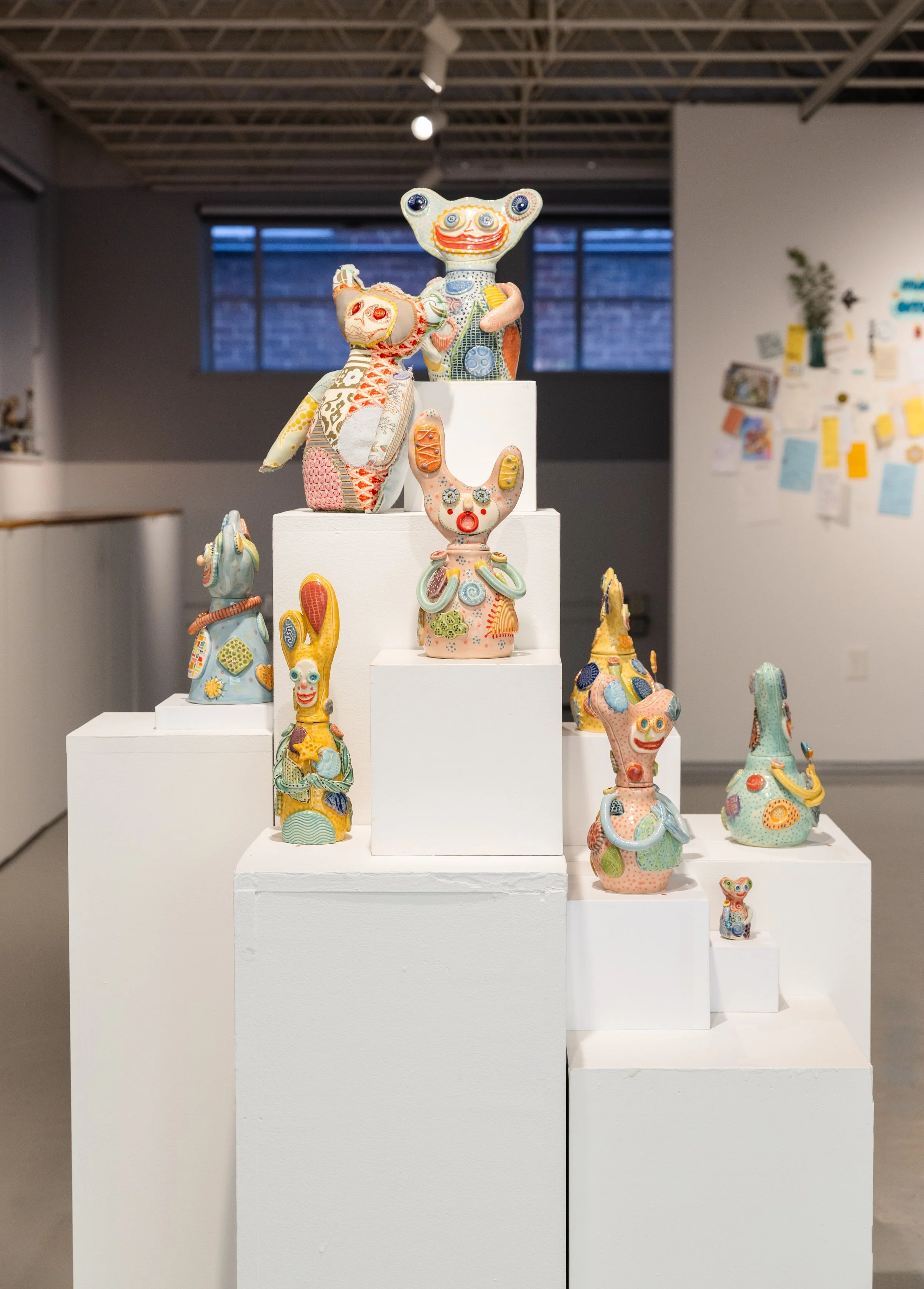 Colorful, hand-decorated animal sculptures stand on white pedestals in a modern gallery, creating a playful, whimsical exhibition against a gray wall.