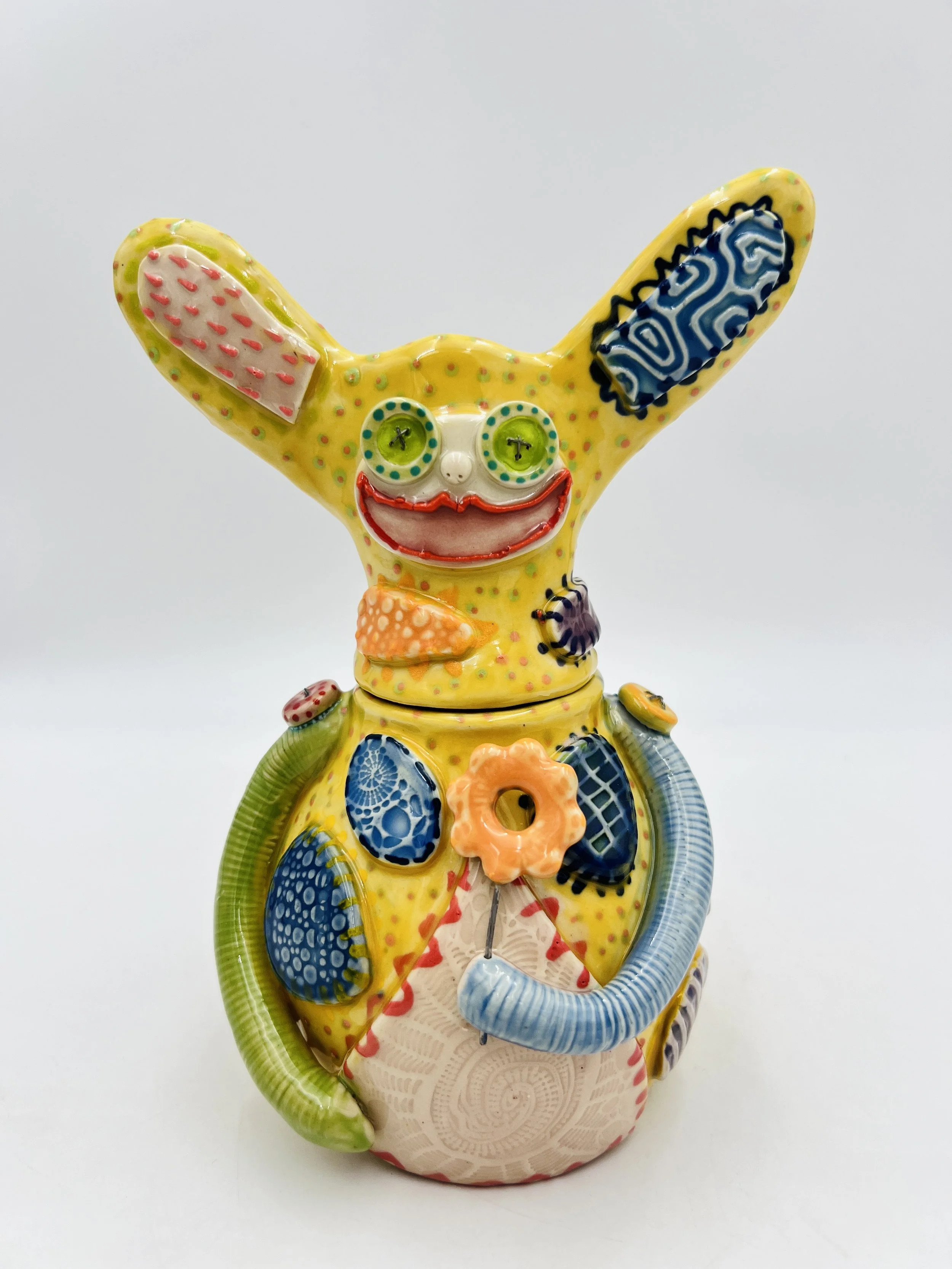 Colorful, whimsical ceramic figures called Gimmies with exaggerated facial features and various patterns, set against a gradient pastel background.