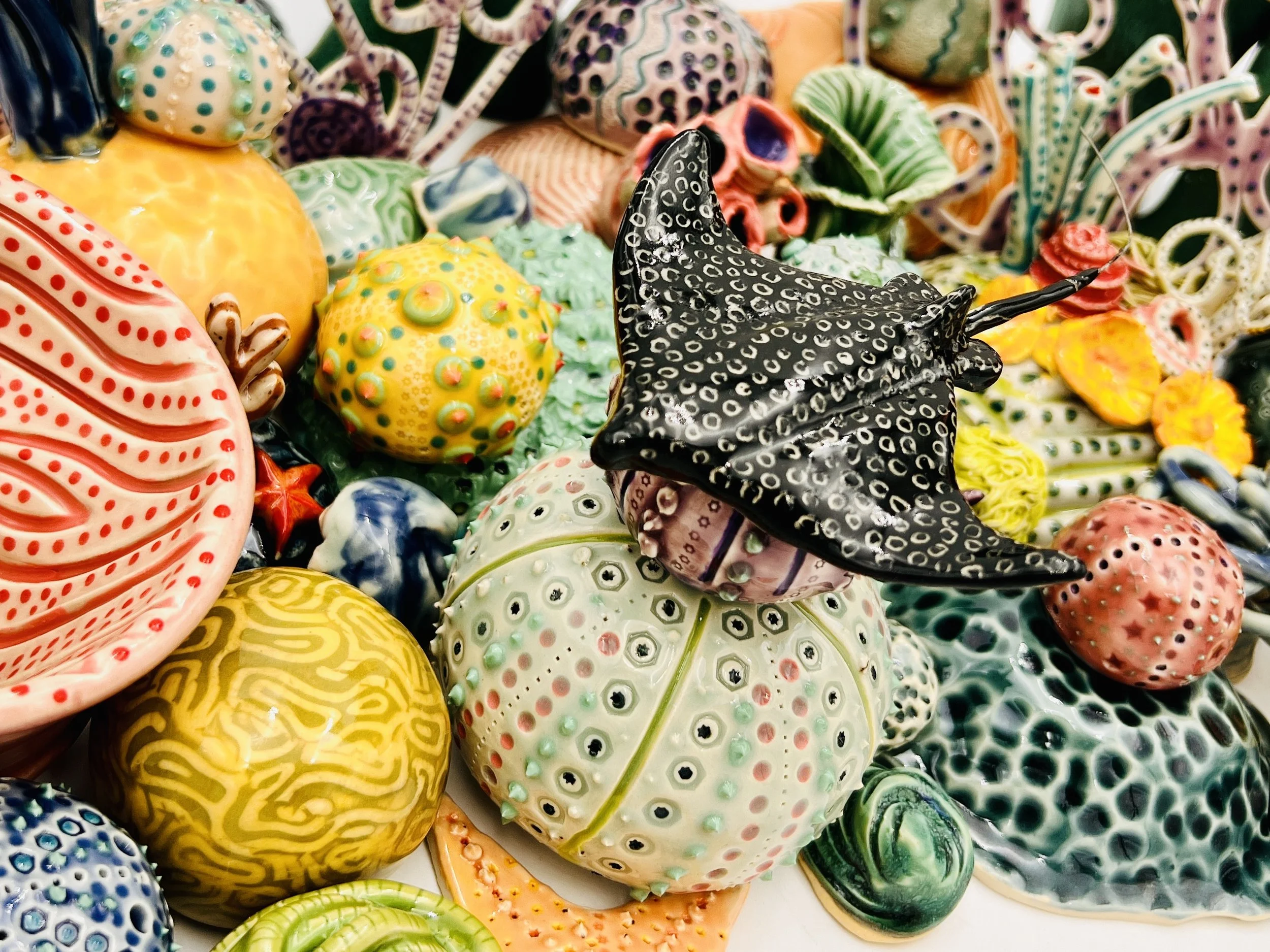 Colorful, textured clay sculptures resembling coral reefs arranged on a gradient background. Each piece has vibrant, intricate details, evoking a sense of ocean life.