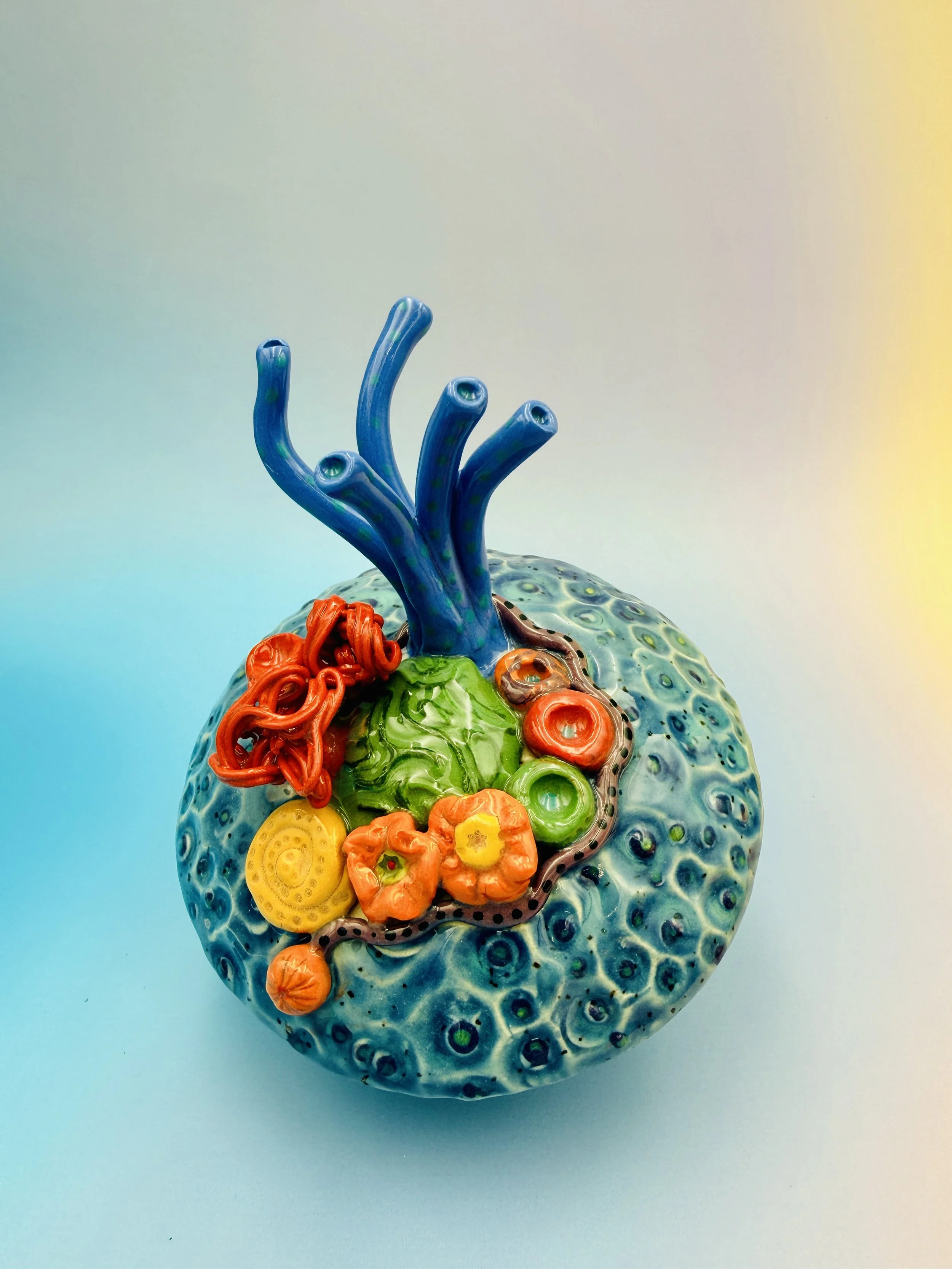 Colorful, textured clay sculptures resembling coral reefs arranged on a gradient background. Each piece has vibrant, intricate details, evoking a sense of ocean life.