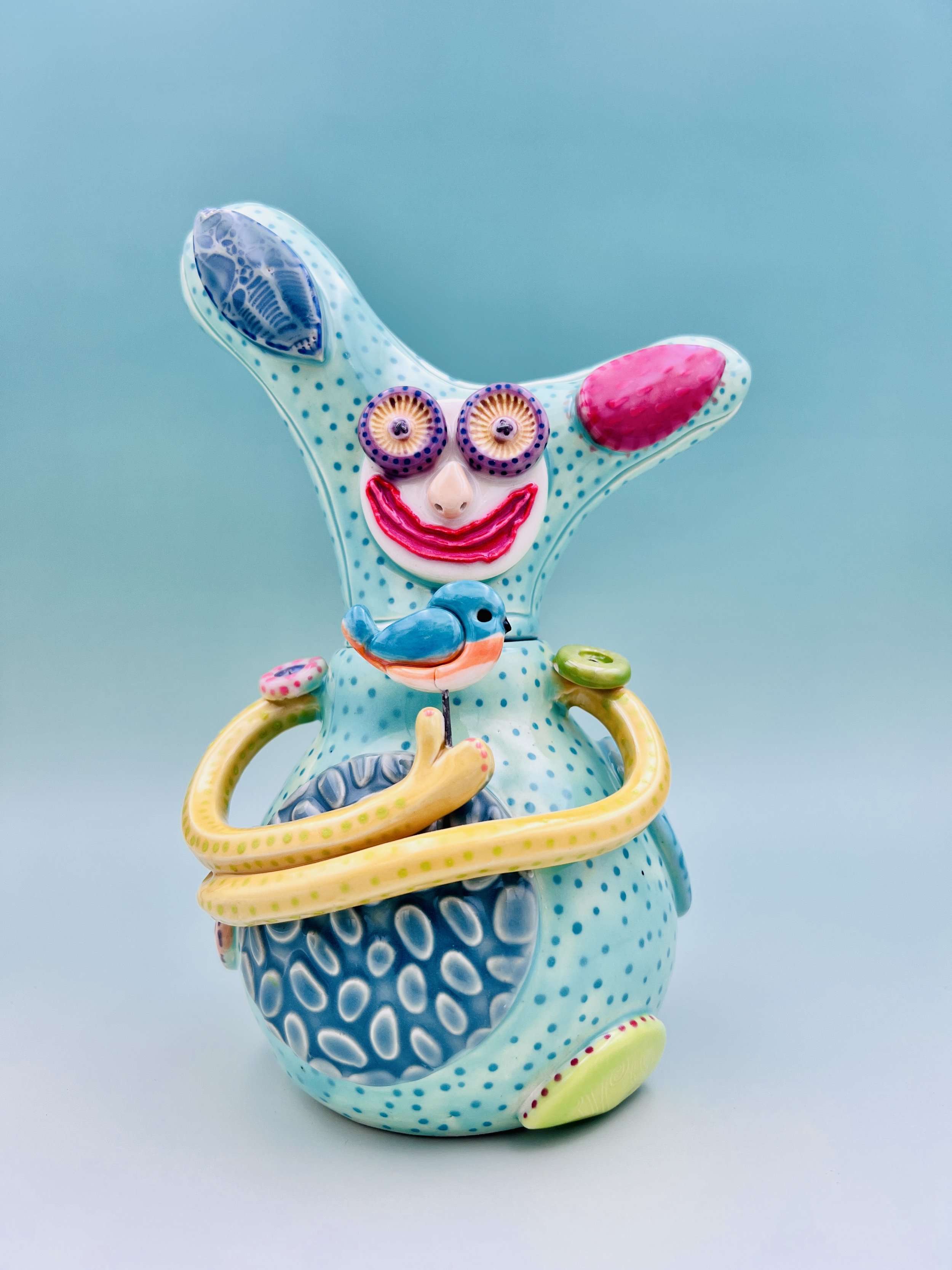 Colorful, whimsical ceramic figures called Gimmies with exaggerated facial features and various patterns, set against a gradient pastel background.