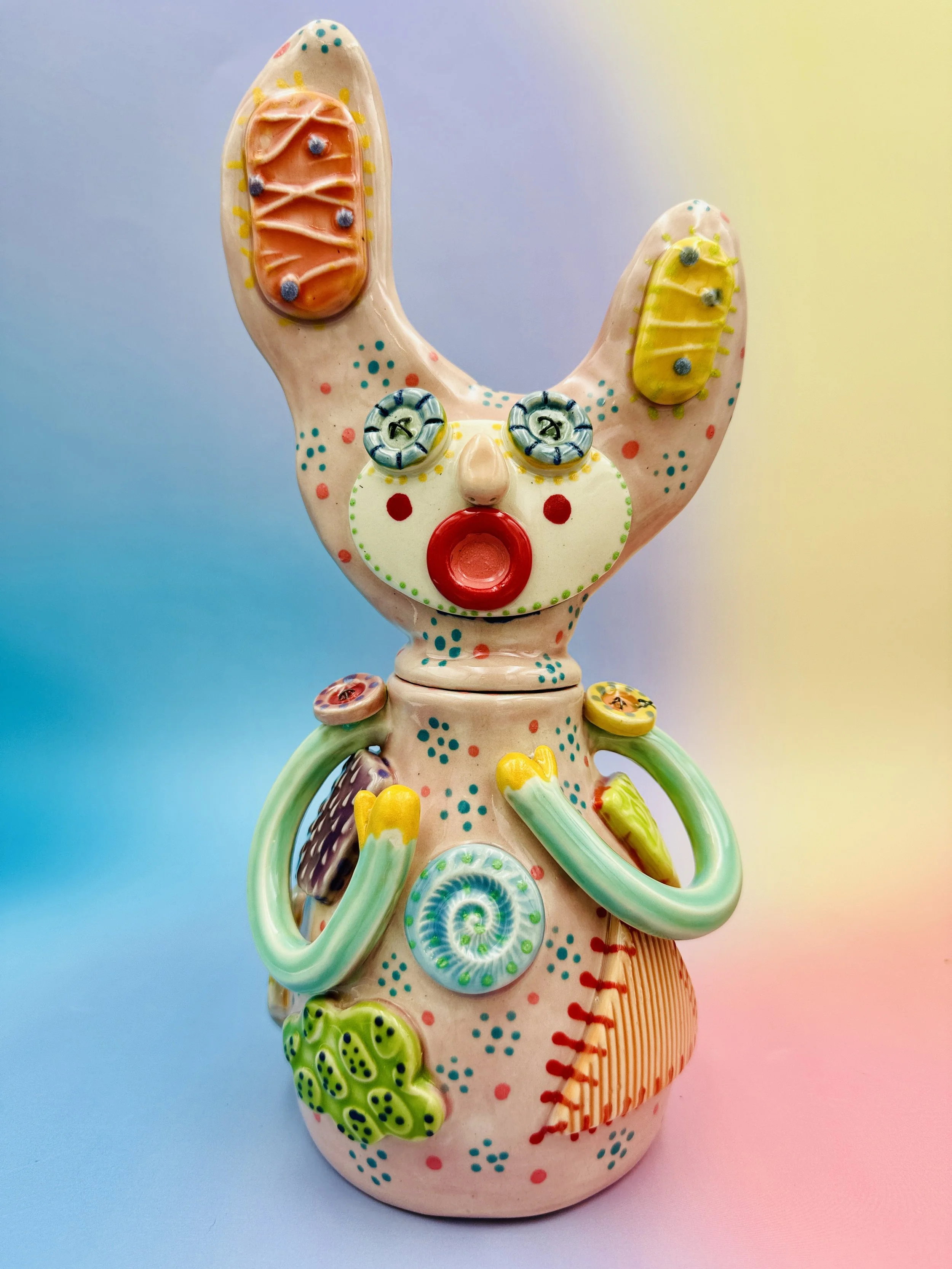 Colorful, whimsical ceramic figures called Gimmies with exaggerated facial features and various patterns, set against a gradient pastel background.