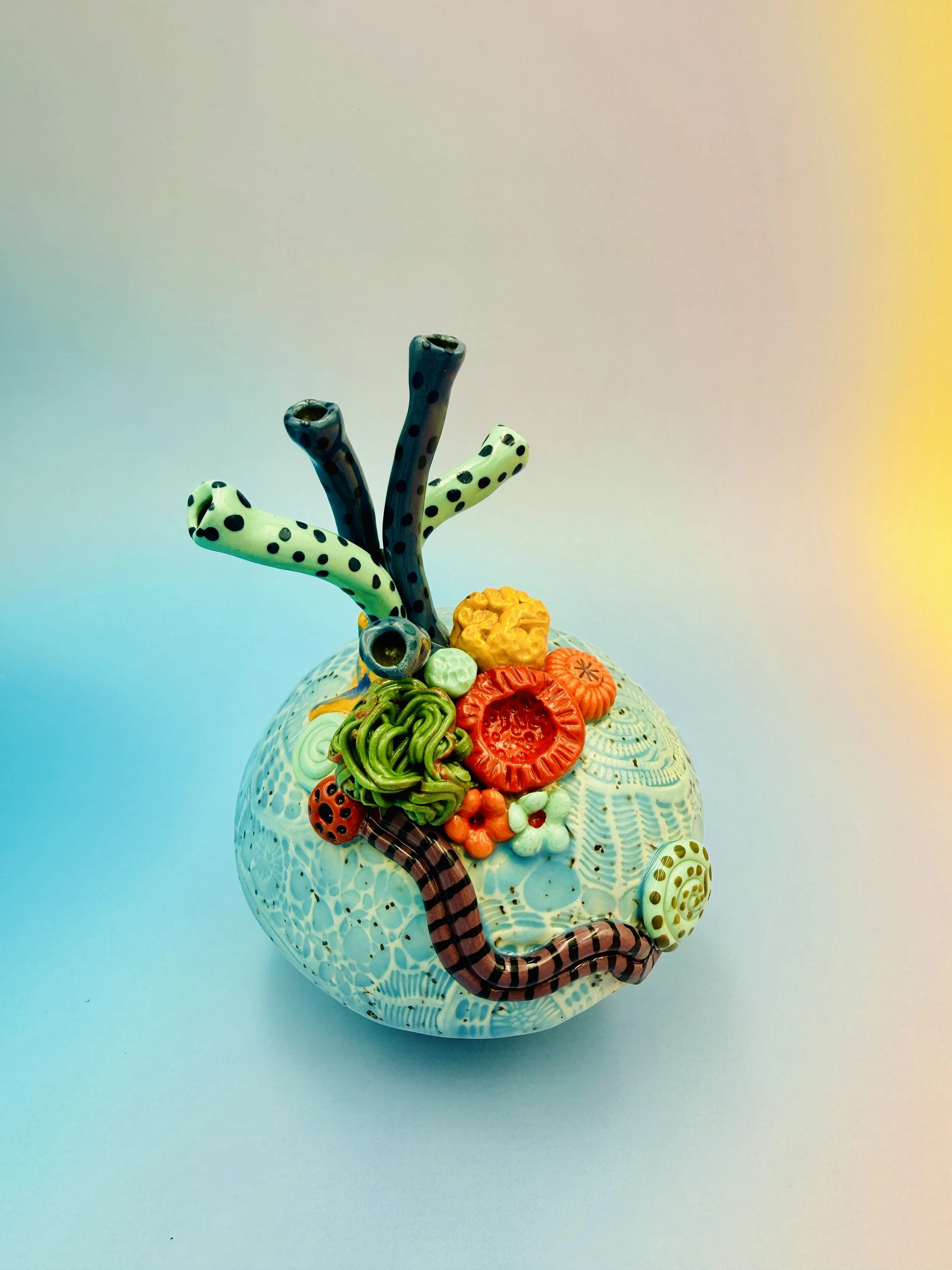 Colorful, textured clay sculptures resembling coral reefs arranged on a gradient background. Each piece has vibrant, intricate details, evoking a sense of ocean life.
