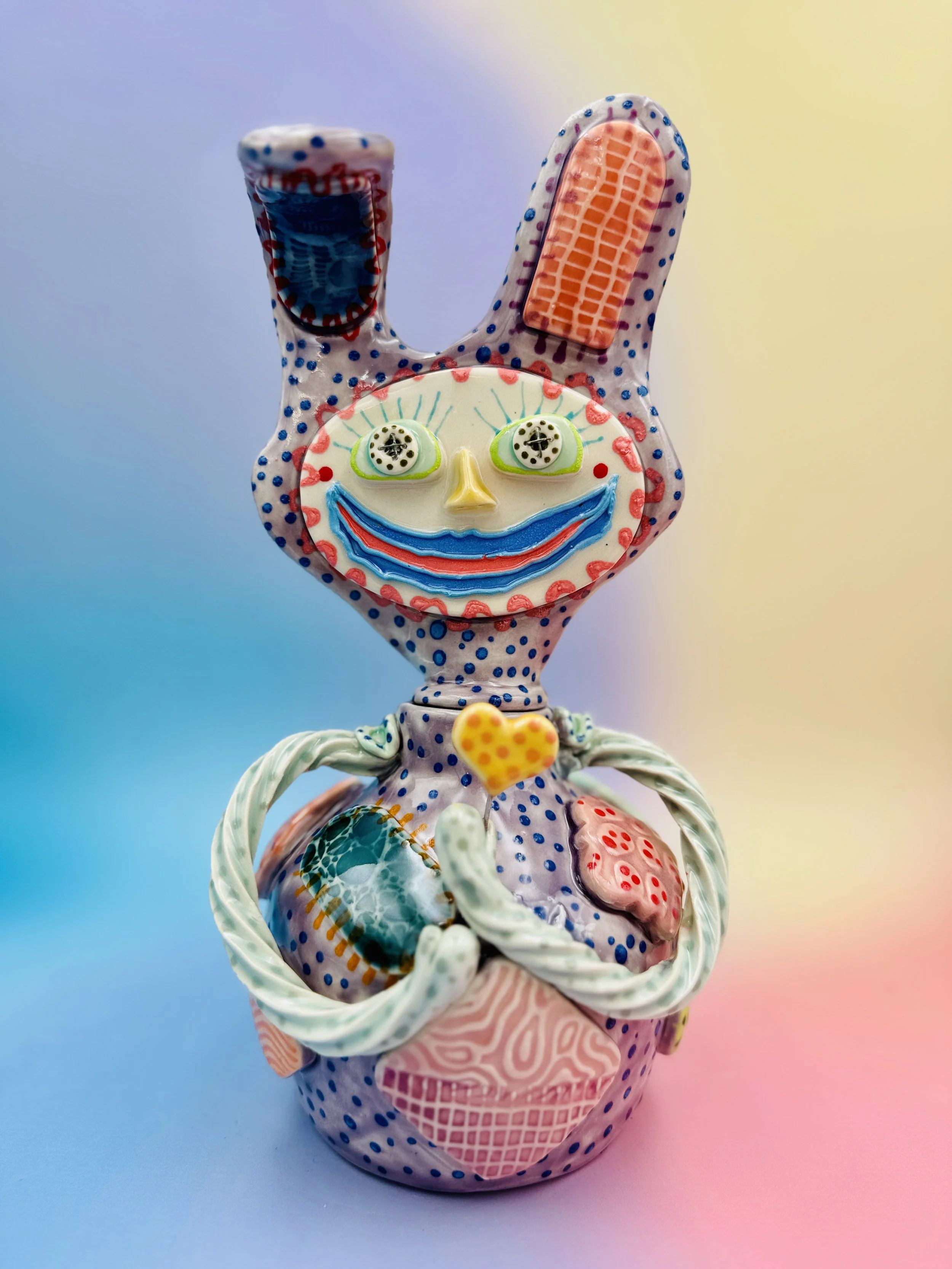 Colorful, whimsical ceramic figures called Gimmies with exaggerated facial features and various patterns, set against a gradient pastel background.