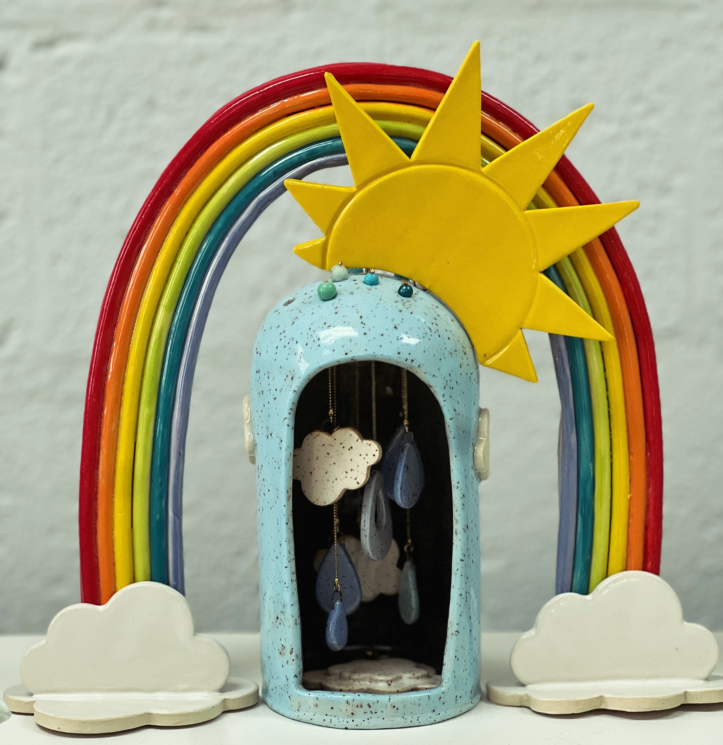 Decorative rainbow with rainbow-colored arches, topped with a yellow sun made of wood, with clouds at the base, and small hanging bells or ornaments underneath.
