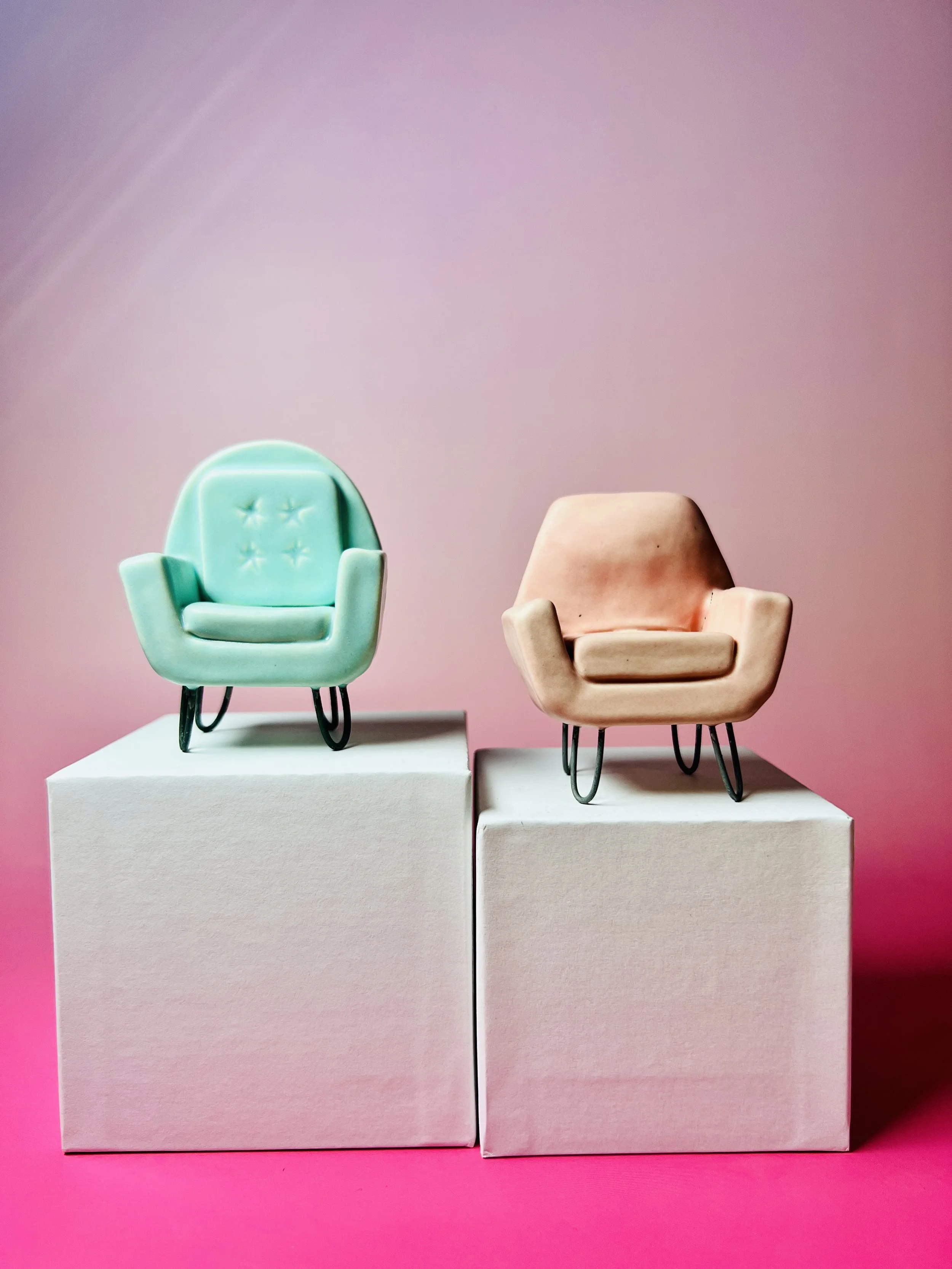  two mid-century modern inspired over stuffed chairs, one turquoise and one pink sitting on top of white platform on a pink background