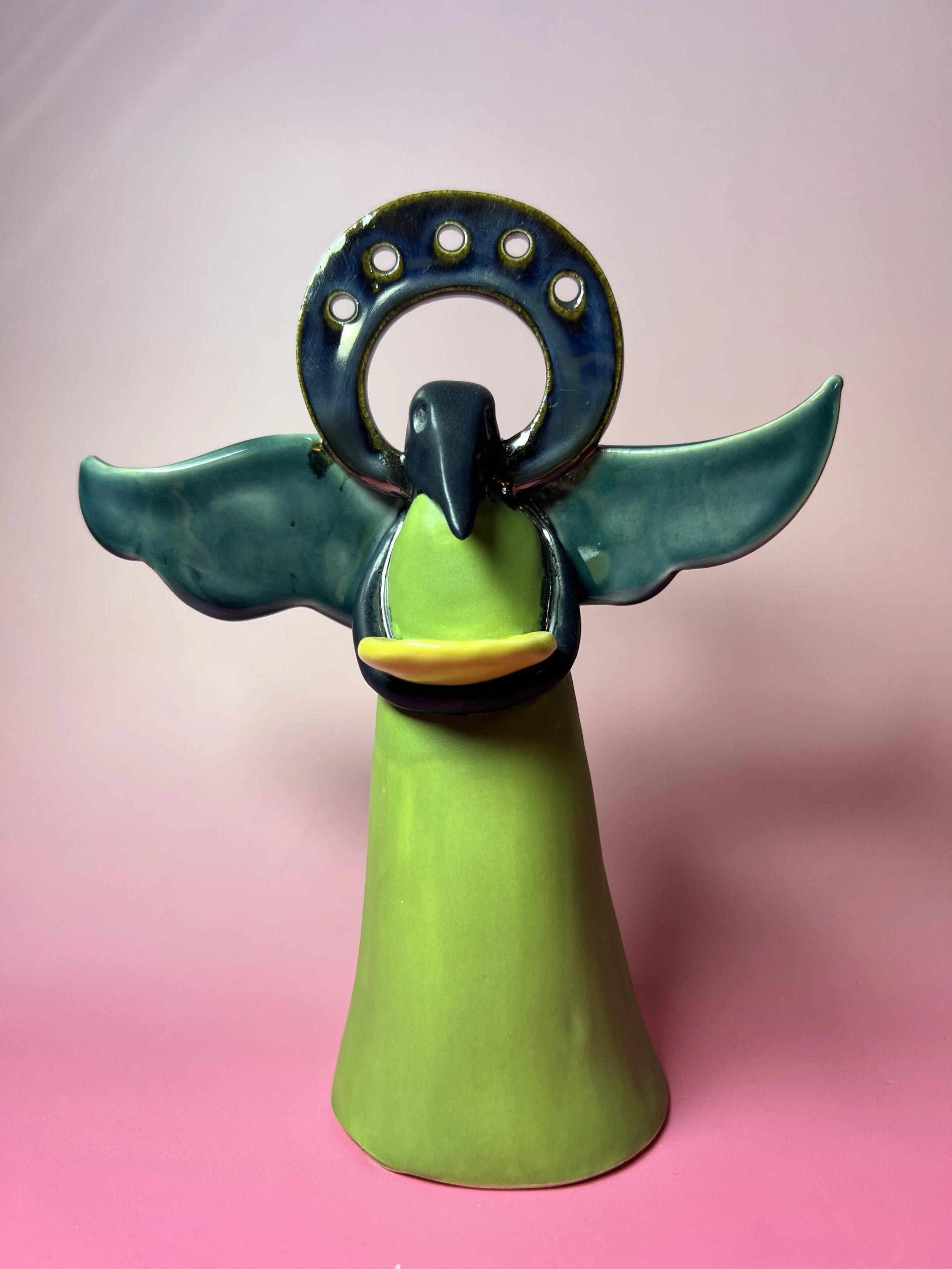 A decorative item resembling a bird with open wings, a circular metal piece above its head, and a green cone-shaped base, set against a pink background.