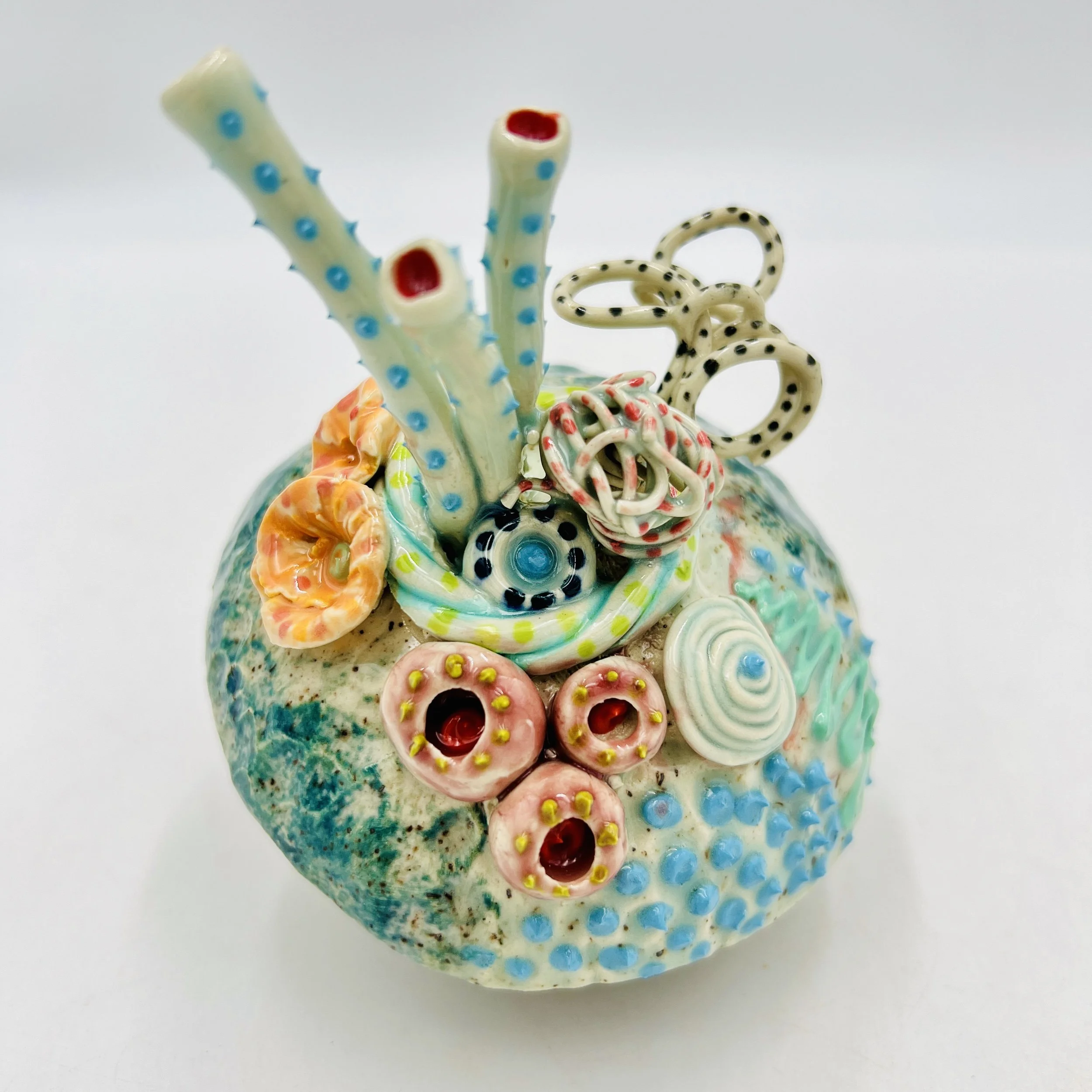 Colorful, textured clay sculptures resembling coral reefs arranged on a gradient background. Each piece has vibrant, intricate details, evoking a sense of ocean life.