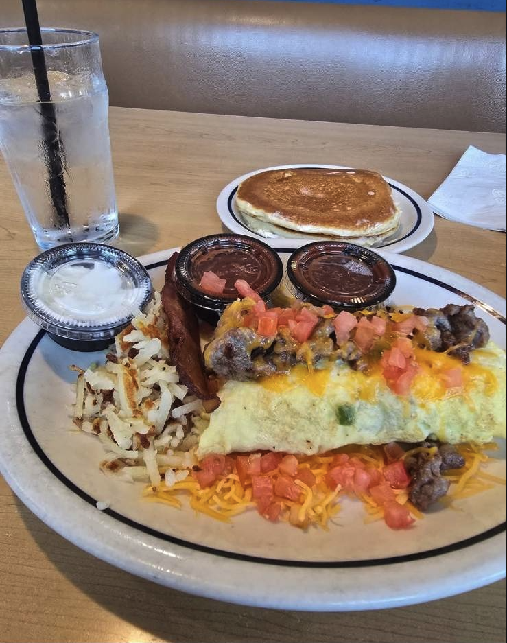 A breakfast plate with scrambled eggs topped with tomatoes and sausage, shredded cheese, bacon, shredded hash browns, and three containers of salsa or sauce. In the background, there are pancakes on a separate plate, a glass of water with a straw, an