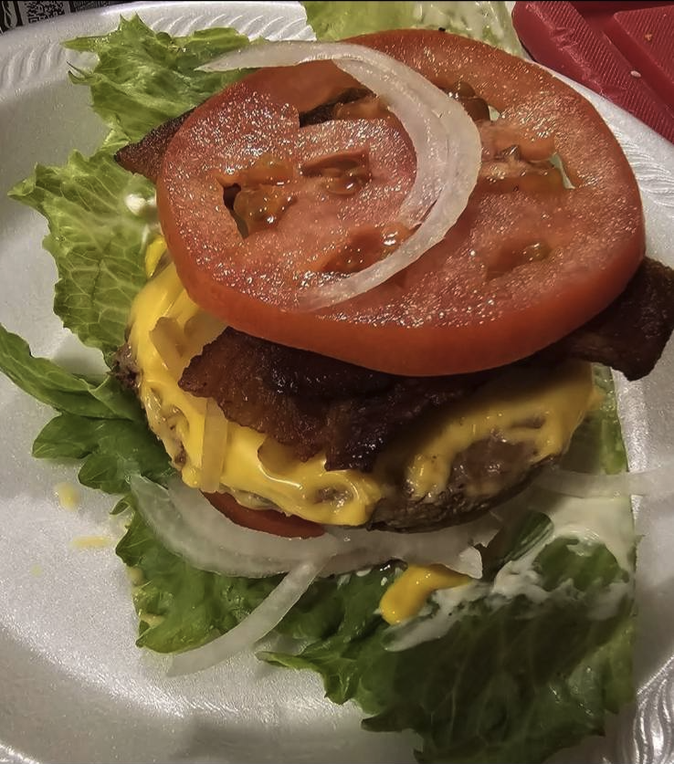 Close-up of an unassembled cheeseburger with lettuce, tomato, onion, bacon, cheese, and beef patty on a foam plate.