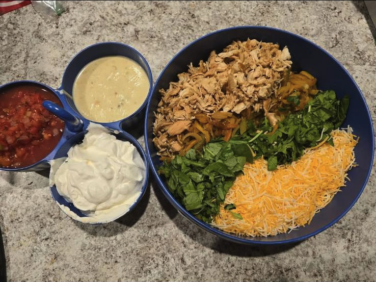 Large bowl with shredded cheese, chopped collard greens, cooked shredded chicken, and sautéed yellow bell peppers. Smaller bowls containing salsa, green queso dip, and sour cream are placed beside the large bowl on a gray countertop.