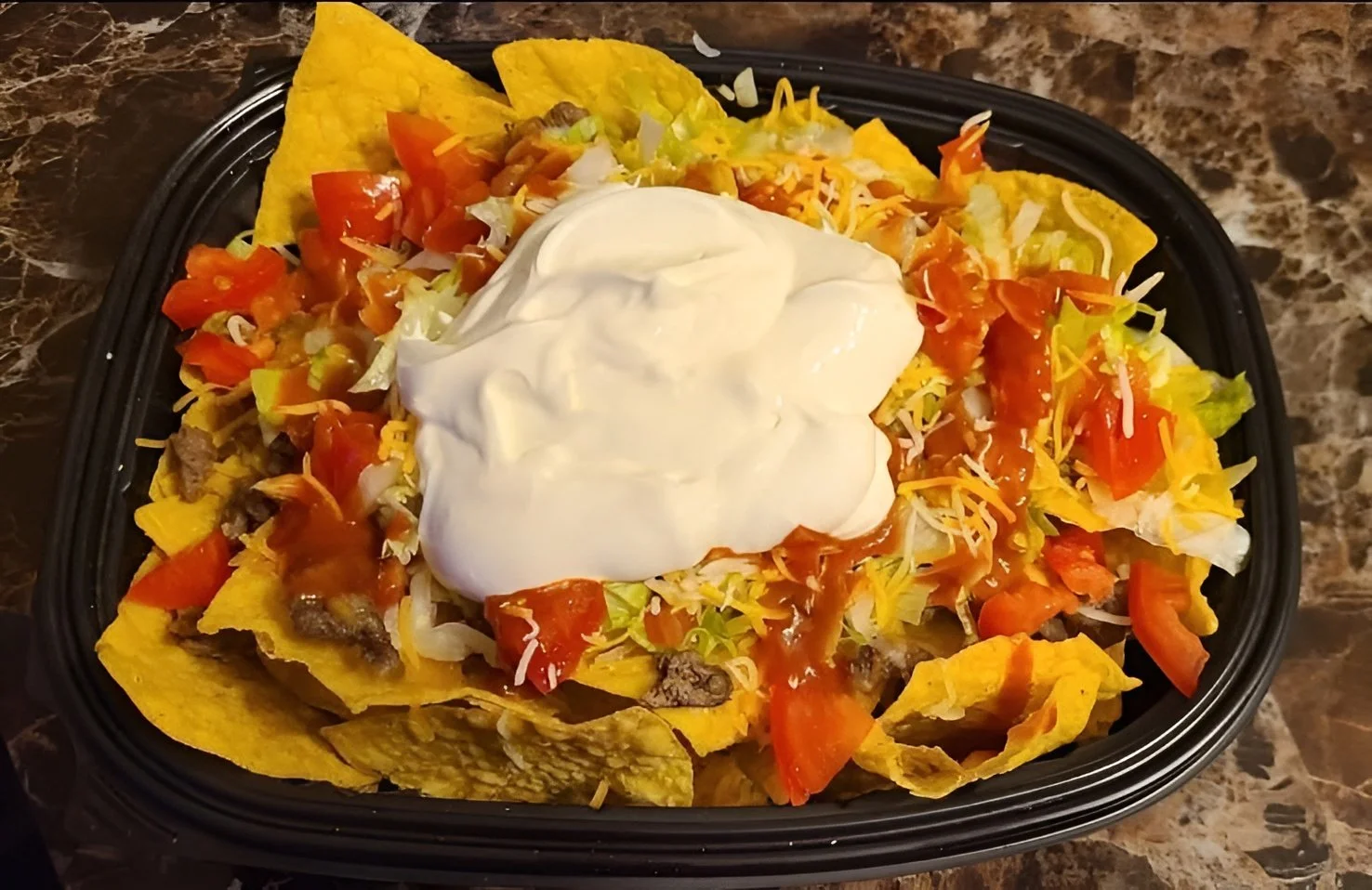 A black plastic tray of nachos with ground beef, lettuce, diced tomatoes, shredded cheese, sour cream, and tortilla chips, on a marble surface.