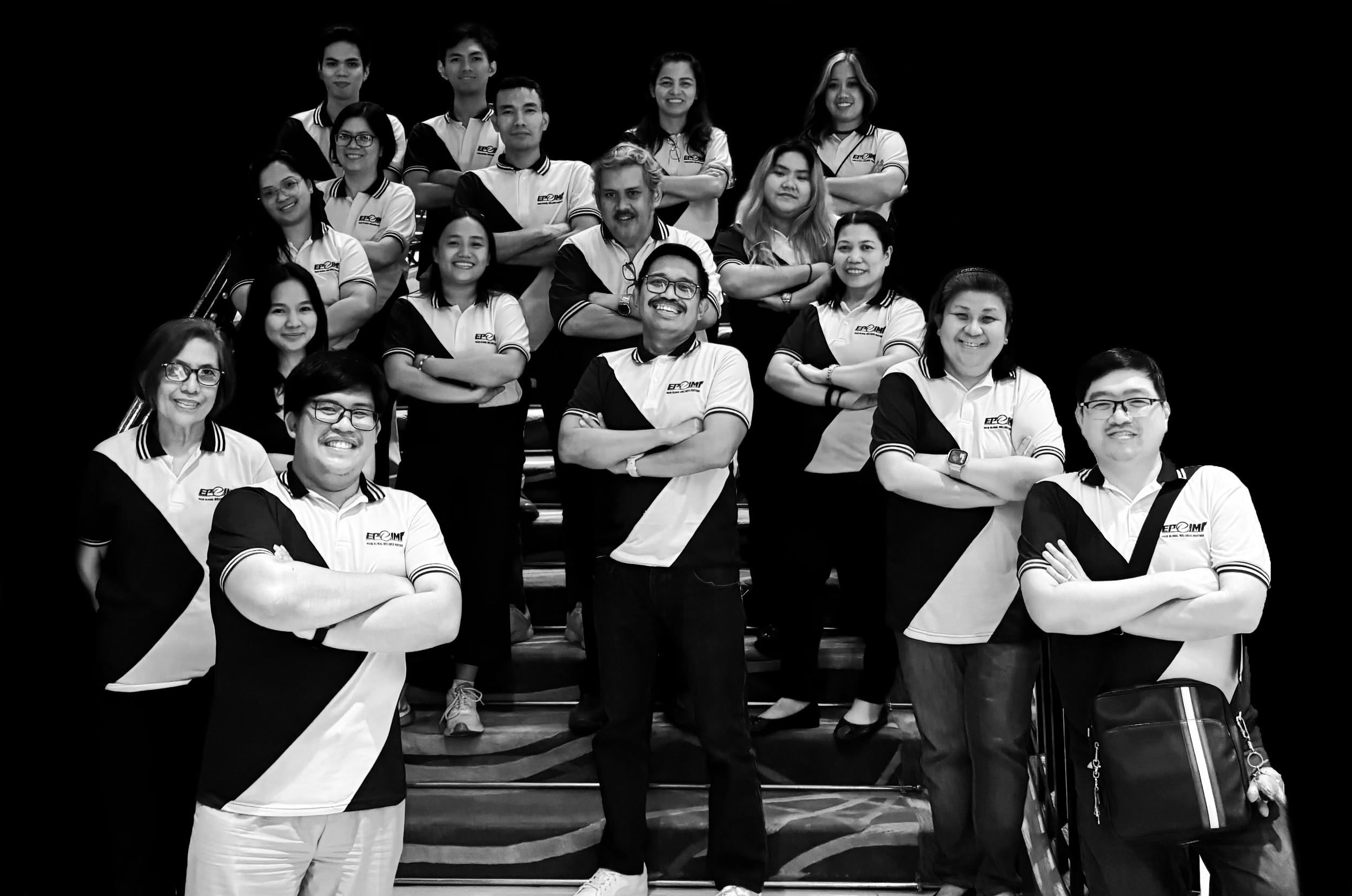 Group portrait of 16 people, standing and sitting on stairs, all wearing matching white and black polo shirts with logo, smiling, in black and white photo.