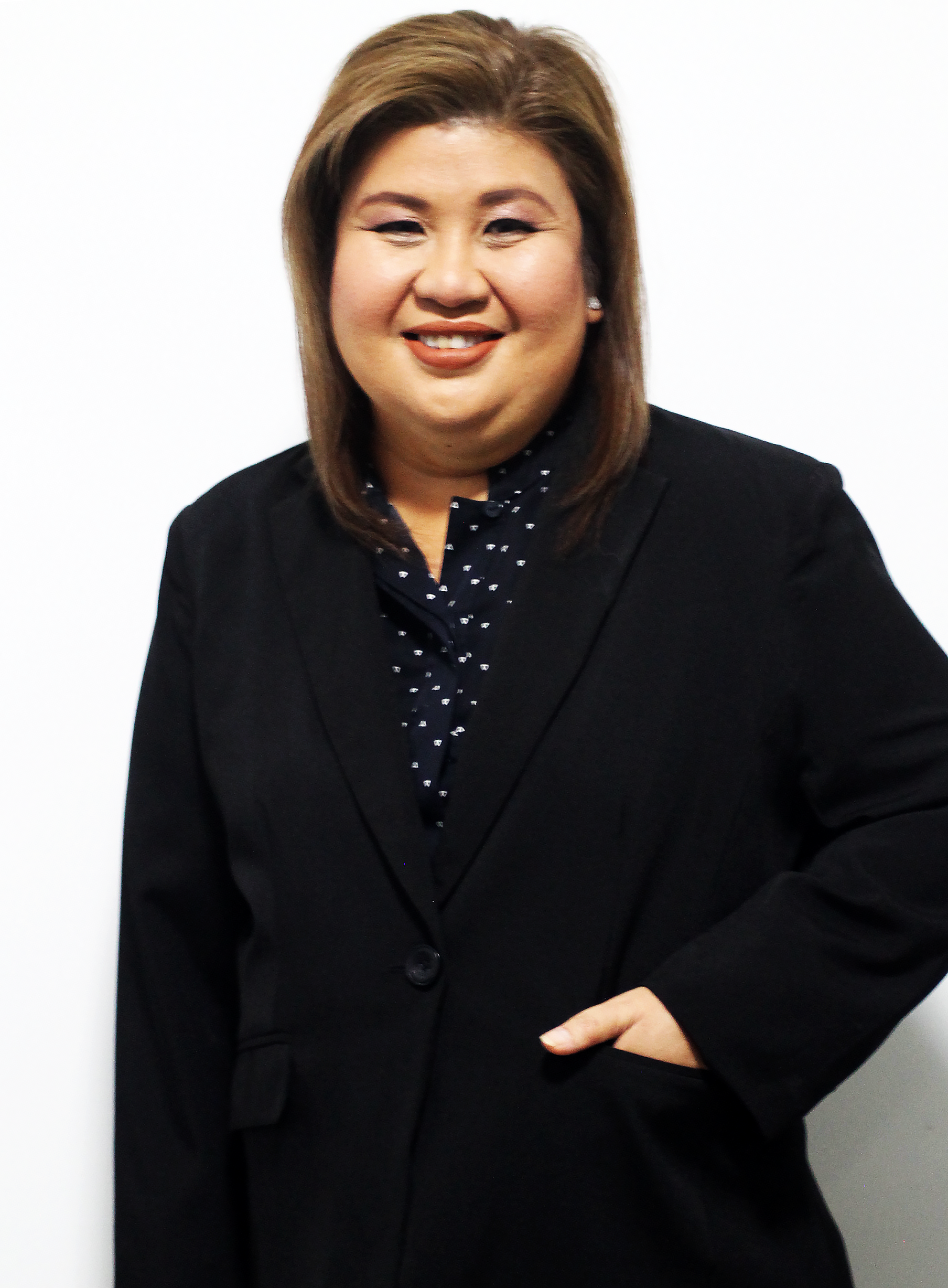 Professional woman with light skin and brown hair, wearing a black blazer and patterned shirt, standing against a white background.