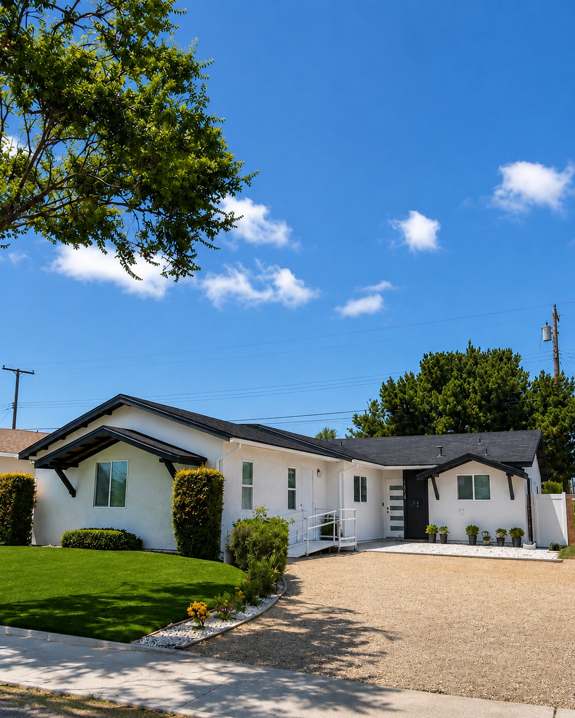 Modern white house with black roof, small front yard with green lawn and potted plants, clear blue sky, trees, and utility poles in the background.