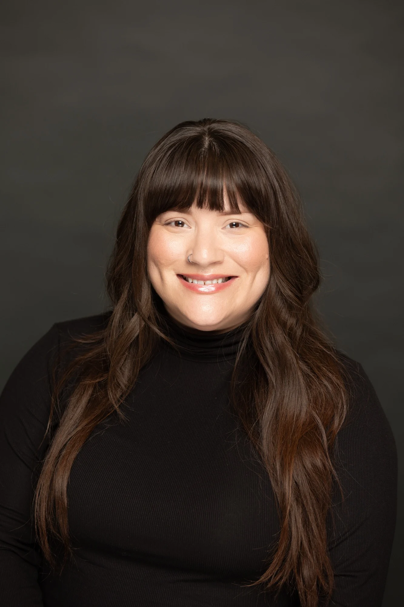 Portrait of a woman with long wavy brown hair, wearing a black turtleneck, smiling against a dark gray background.