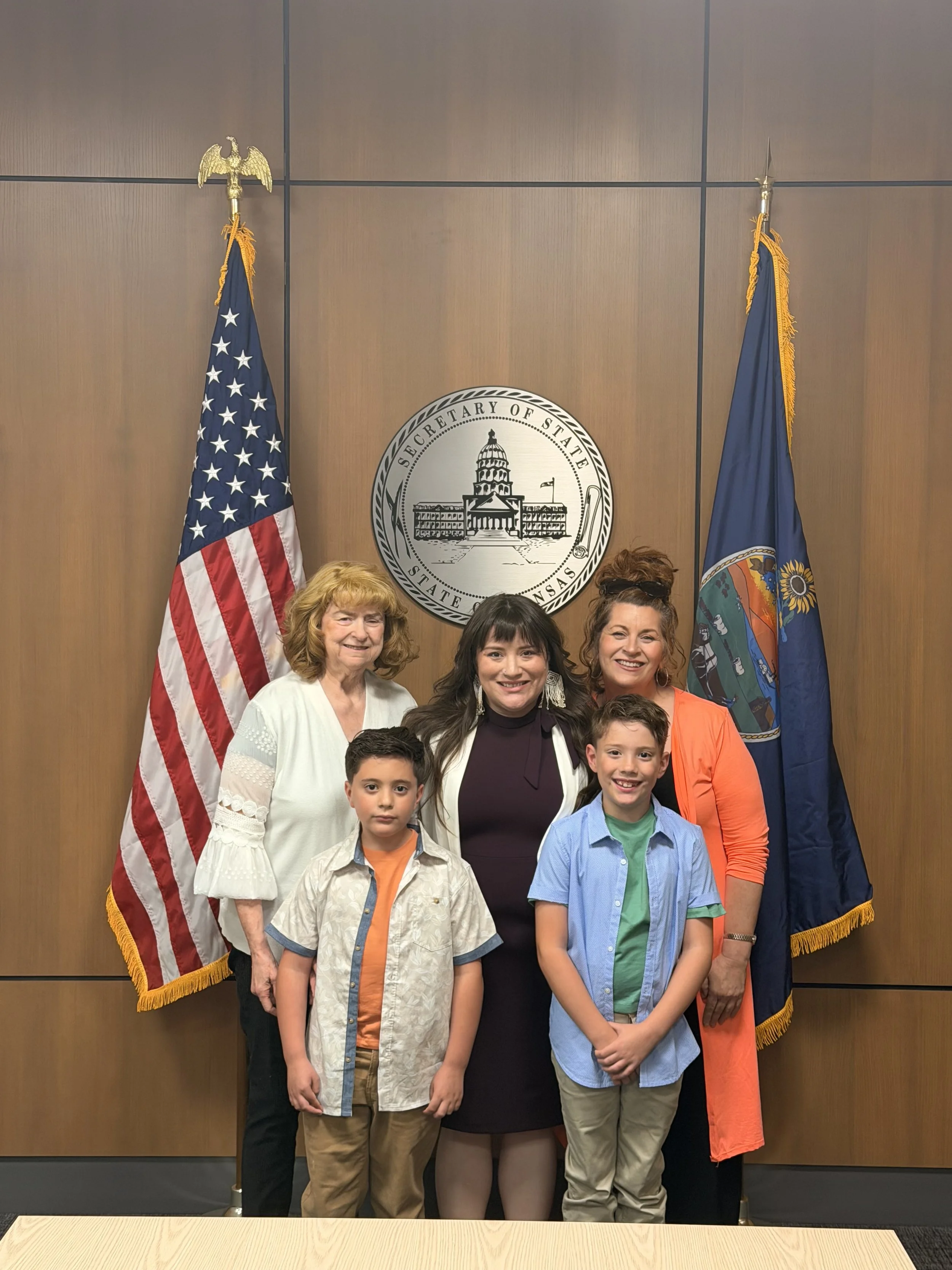 Group of seven people, including children and adults, standing in front of a wooden wall with U.S. and state flags, and a round seal that reads 'Secretary of State Arkansas'.