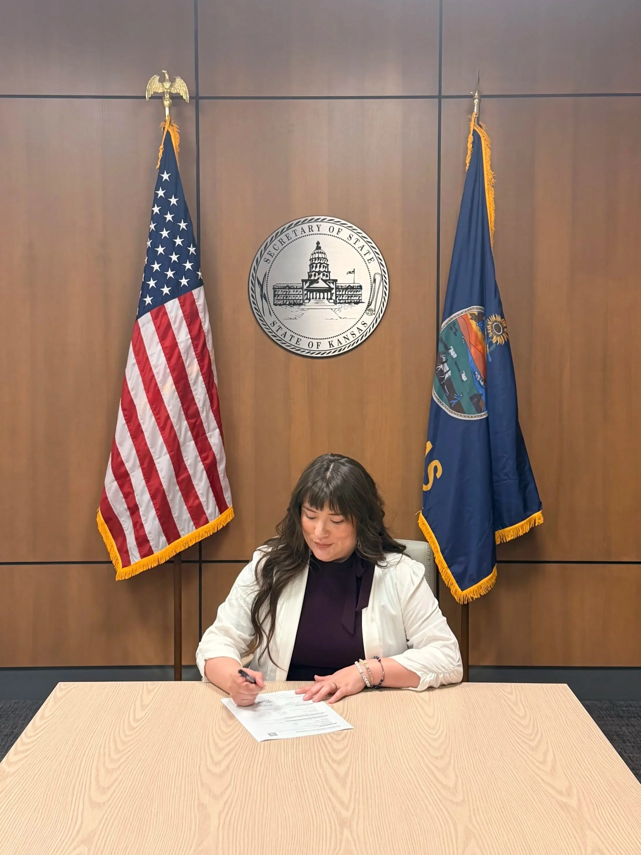 A woman signing a document at a wooden table in an office setting with two flags and a state seal of Kansas behind her.